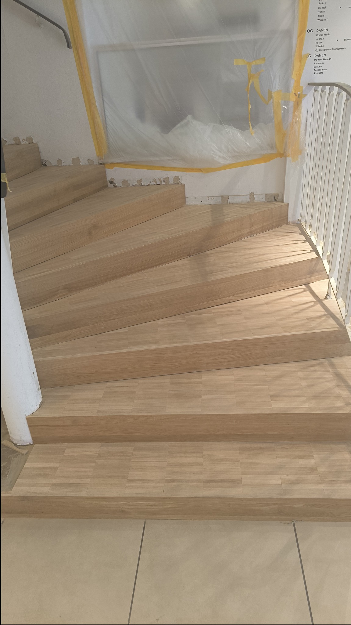 Schody wyłożone panelami drewnopodobnymi w trakcie prac wykończeniowych; zabezpieczone folią okno i fragment ściany z widocznym napisem 'DAMEN'.