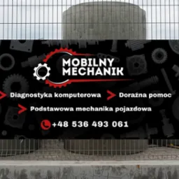 Baner reklamowy z napisem 'Mobilny Mechanik' i numerem telefonu, umieszczony na metalowym ogrodzeniu na tle betonowych kręgów i pomarańczowych rur.