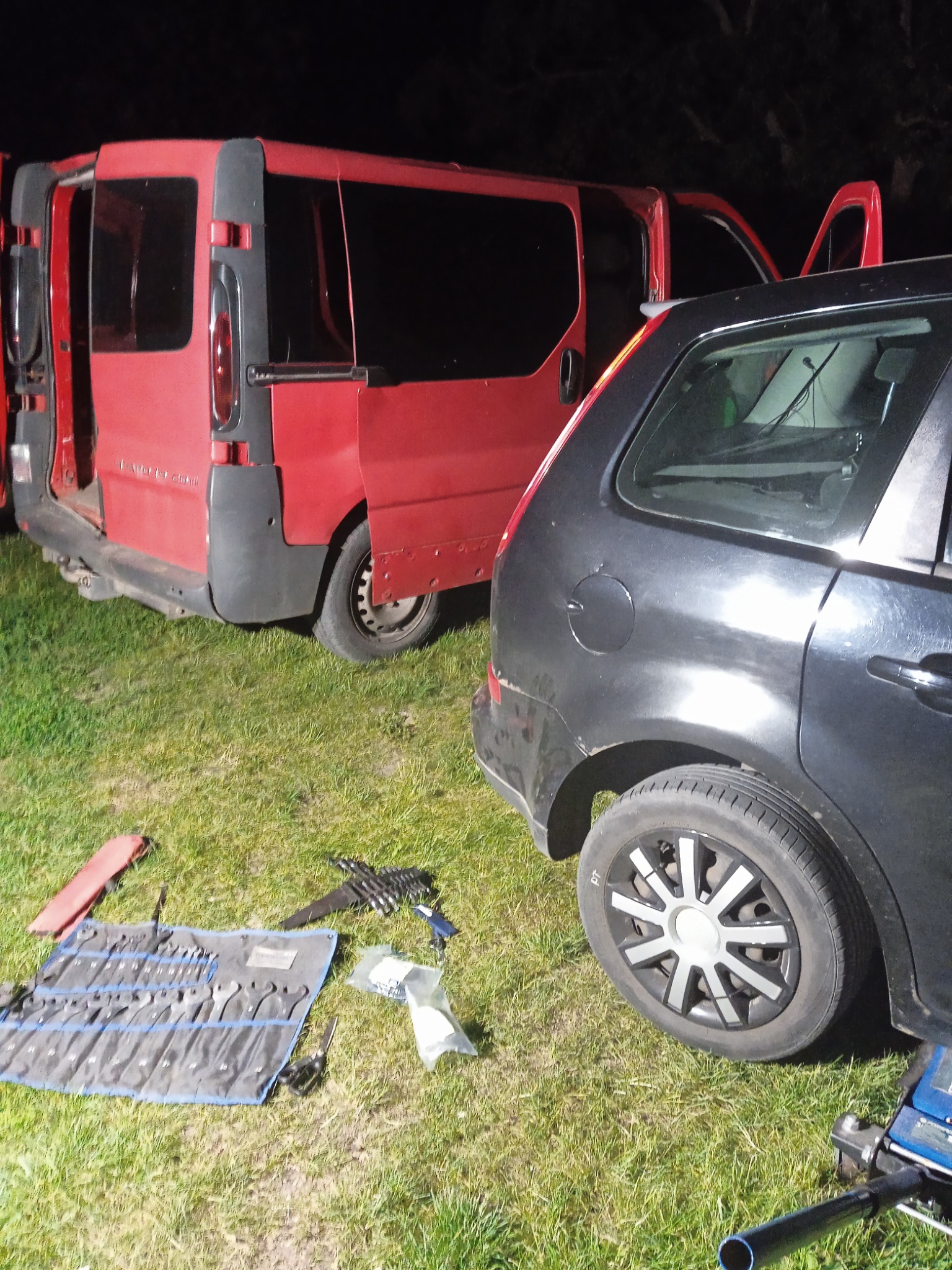 Dwa samochody na trawie, czerwony bus z otwartymi drzwiami i czarny hatchback, obok rozłożony zestaw kluczy i podnośnik hydrauliczny.