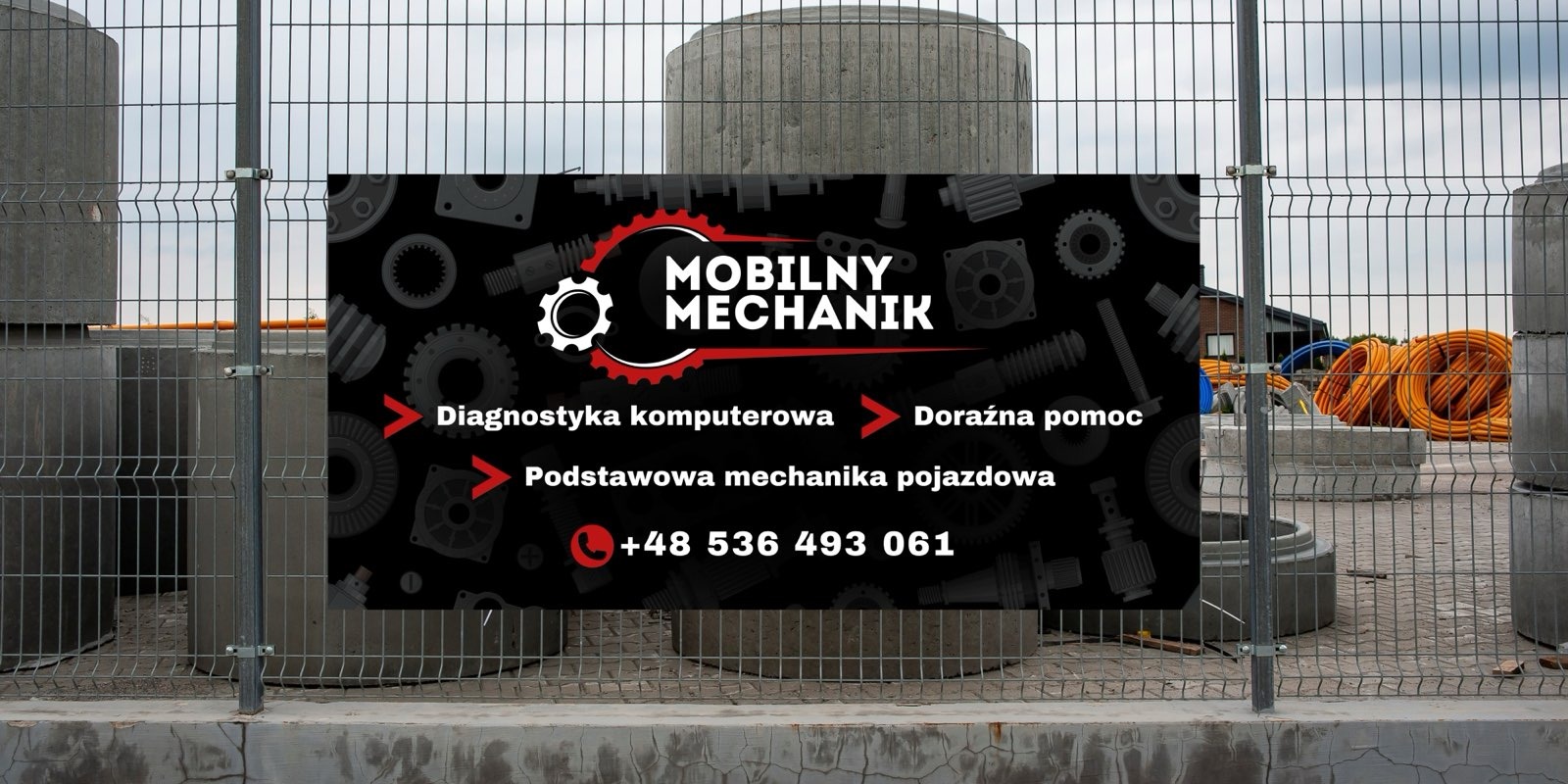 Baner reklamowy z napisem 'Mobilny Mechanik' i numerem telefonu, umieszczony na metalowym ogrodzeniu na tle betonowych kręgów i pomarańczowych rur.
