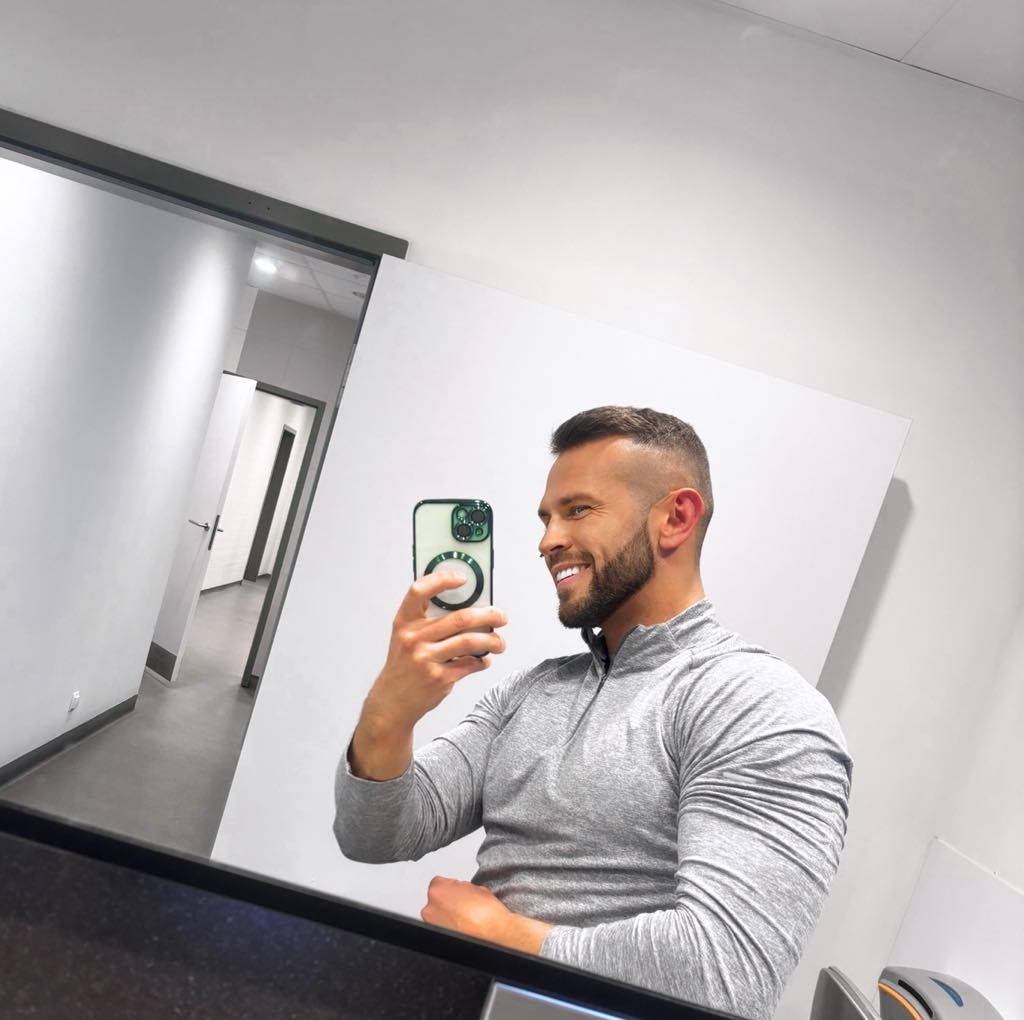 Uśmiechnięty mężczyzna w szarej bluzie robi selfie w lustrze w siłowni, widoczny korytarz z drzwiami w tle.