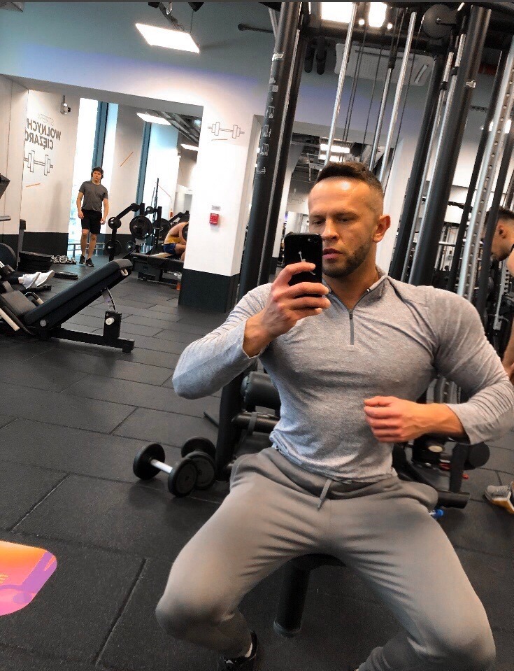 Mężczyzna robi selfie w siłowni, siedząc na maszynie do ćwiczeń; w tle widoczne inne osoby i sprzęt do ćwiczeń.