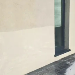 Mycie kostki brukowej przy elewacji budynku z oknem, widoczne ślady wody i piany na czarnej kostce brukowej, kontrast między czystą i brudną częścią elewacji.