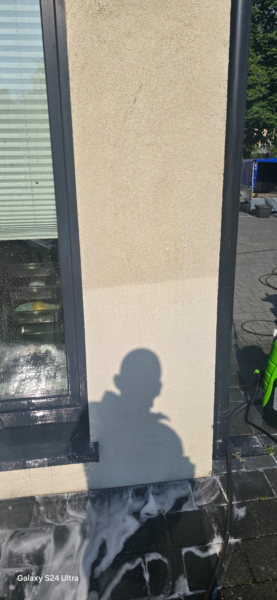 Fragment elewacji budynku z widocznym cieniem osoby podczas czyszczenia. Część ściany jest już umyta, a na kostce brukowej widać pozostałości piany.