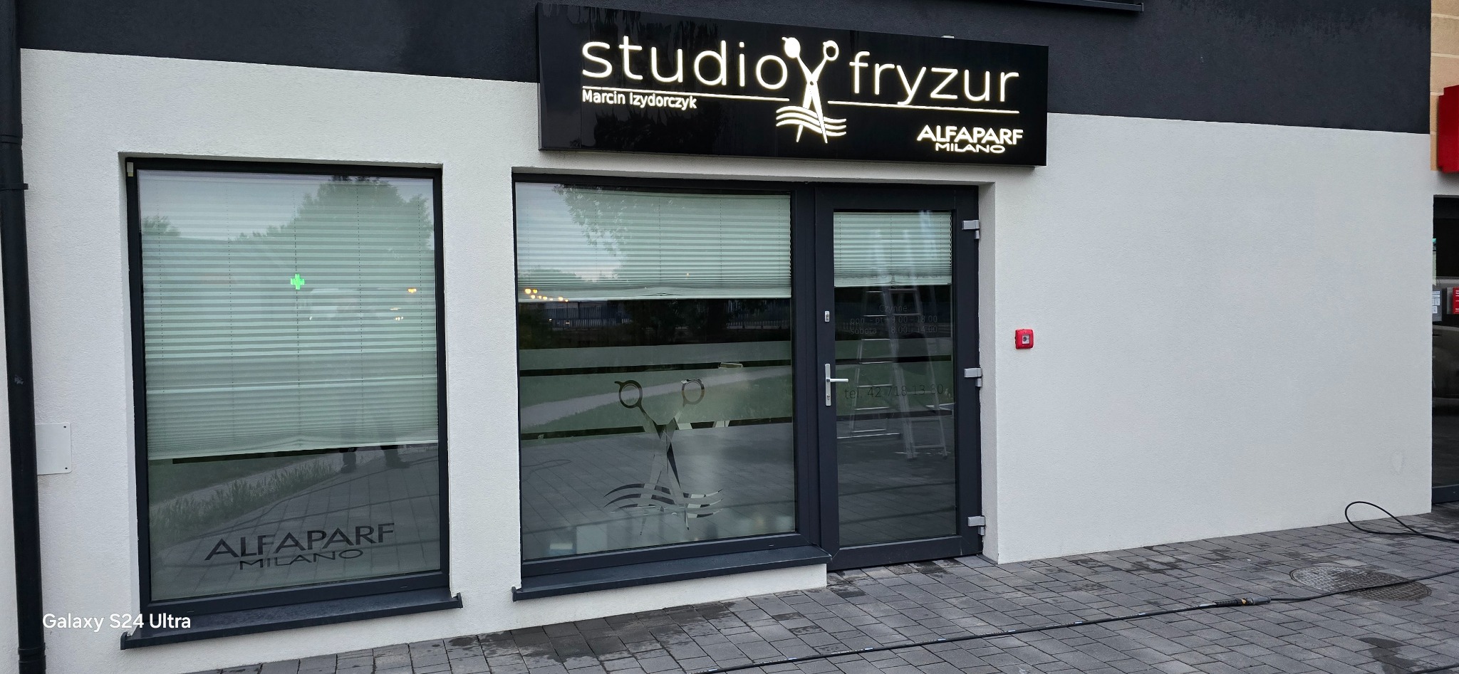 Fasada budynku salonu fryzjerskiego z czarnym szyldem 'studio fryzur Marcin Izydorczyk' i logo Alfaparf Milano, szare drzwi wejściowe z naklejką z grafiką nożyczek i godzinami otwarcia, okna...