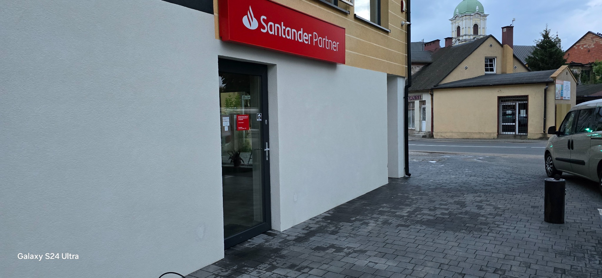 Odnowiona elewacja budynku Santander Partner z gładką, białą ścianą i widocznym logo na czerwonym tle, z ciemną ramą okienną i kostką brukową przed budynkiem.