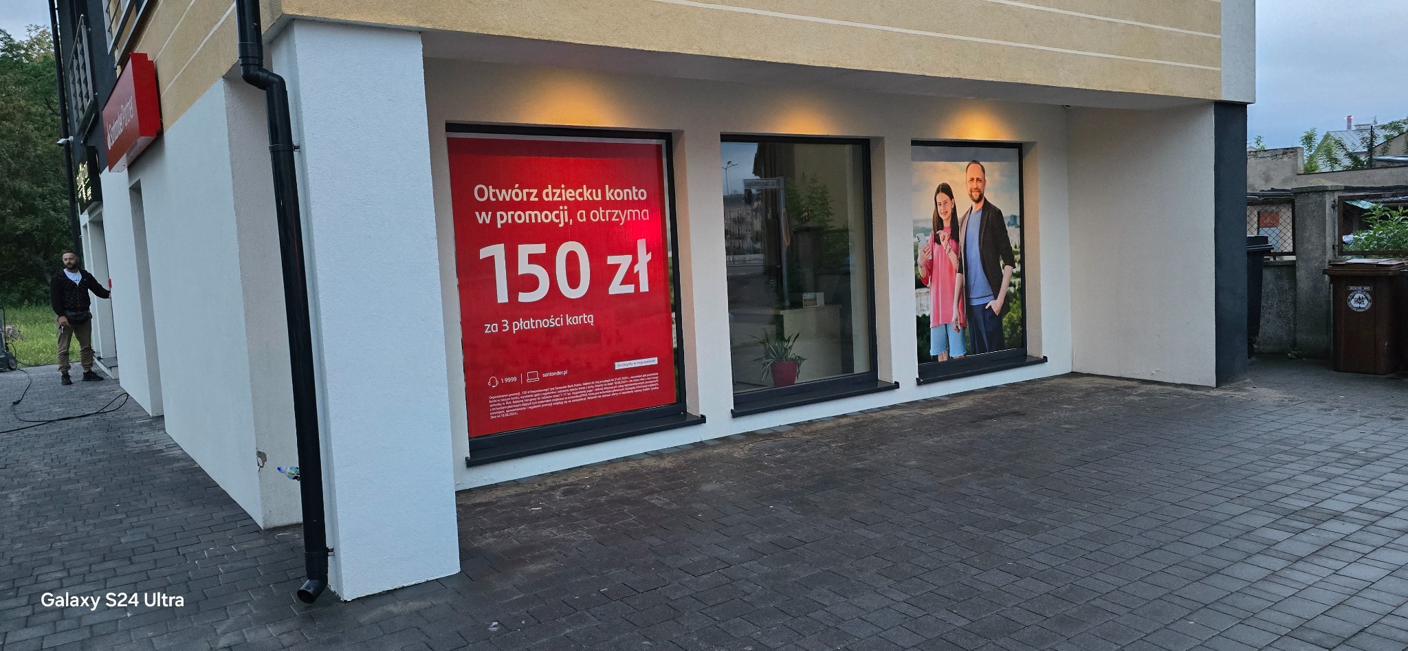 Budynek banku z trzema oknami wystawowymi: na czerwonym plakacie promocja konta dla dzieci, w środkowym oknie roślina doniczkowa, na plakacie w prawym oknie wizerunek uśmiechniętej rodziny.
