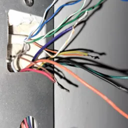 Szczegółowe ujęcie przewodów elektrycznych różnego koloru wychodzących z otworu w ścianie, zamocowanych do metalowej płytki montażowej, z widocznymi końcówkami kabli przygotowanymi do podłączenia.