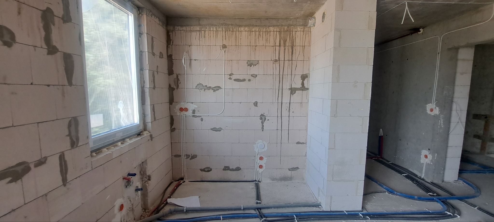 Surowy stan pomieszczenia z widoczną instalacją elektryczną i wodną w ścianach z betonu komórkowego, okno z widokiem na zieleń, niebieskie rury na podłodze.