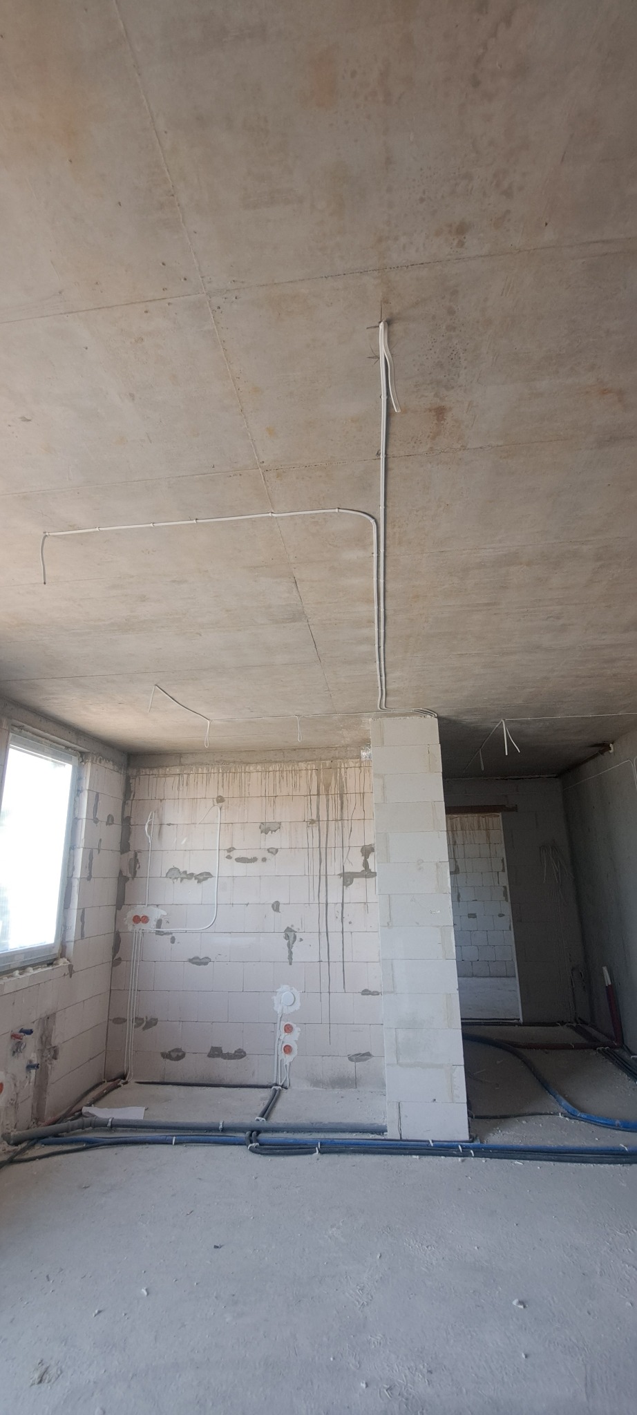 Surowy stan wnętrza z widoczną instalacją elektryczną: białe przewody poprowadzone po suficie i ścianach z betonu komórkowego, widoczne puszki elektryczne, niebieskie rury na podłodze, okno z lewej...