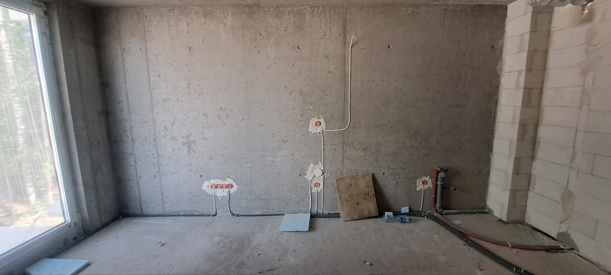 Surowy stan wnętrza z widoczną instalacją elektryczną i hydrauliczną w trakcie montażu, ściany z betonu architektonicznego, widoczne puszki elektryczne i rury wodne, częściowo zakryte płytą...