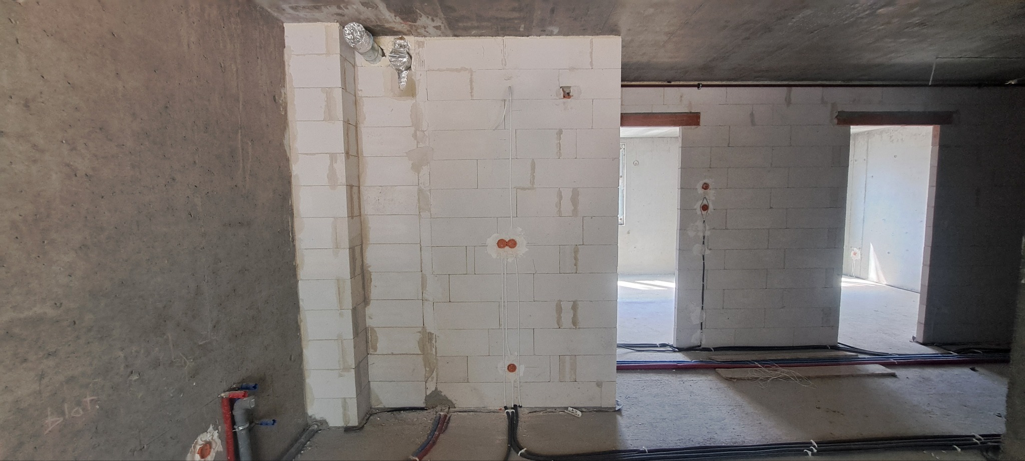 Wnętrze w stanie deweloperskim z widocznymi przewodami elektrycznymi w ścianach z betonu komórkowego oraz rurami instalacji wodnej przy ścianie z surowym betonem, widoczna instalacja wentylacyjna...