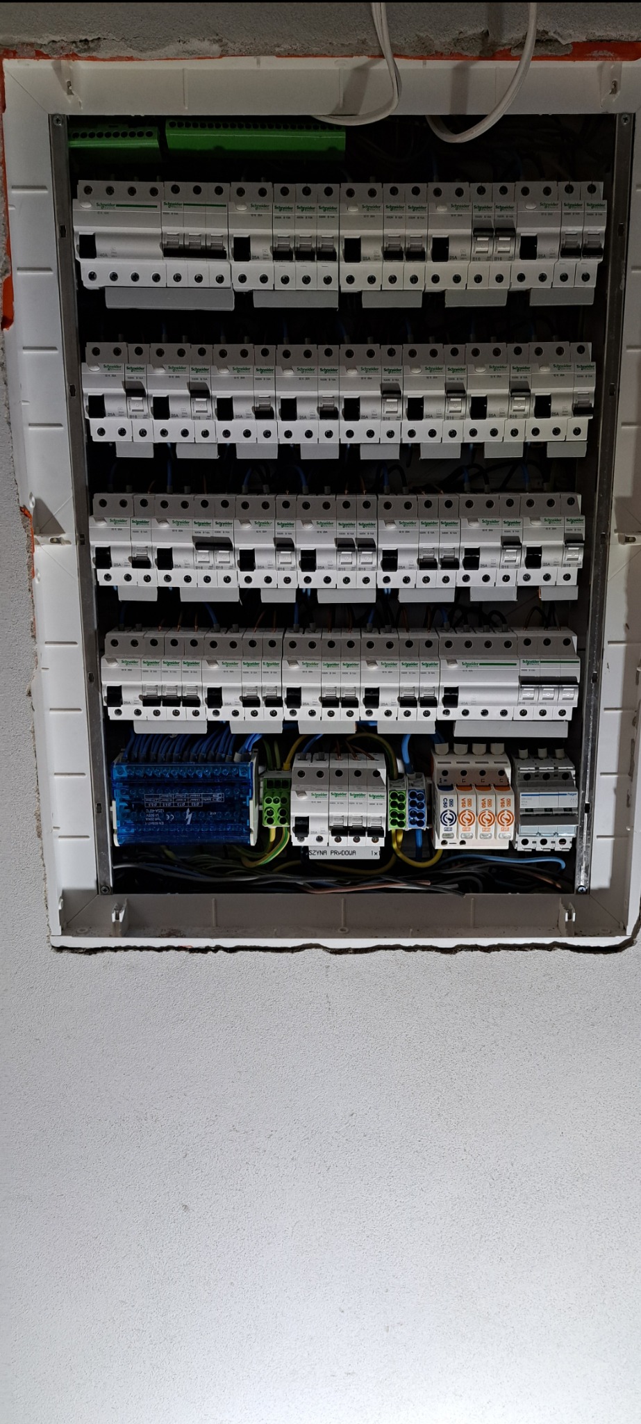 Otwarta skrzynka rozdzielcza z licznymi bezpiecznikami i przewodami, widoczne detale instalacji elektrycznej wewnątrz, w tle surowa ściana.
