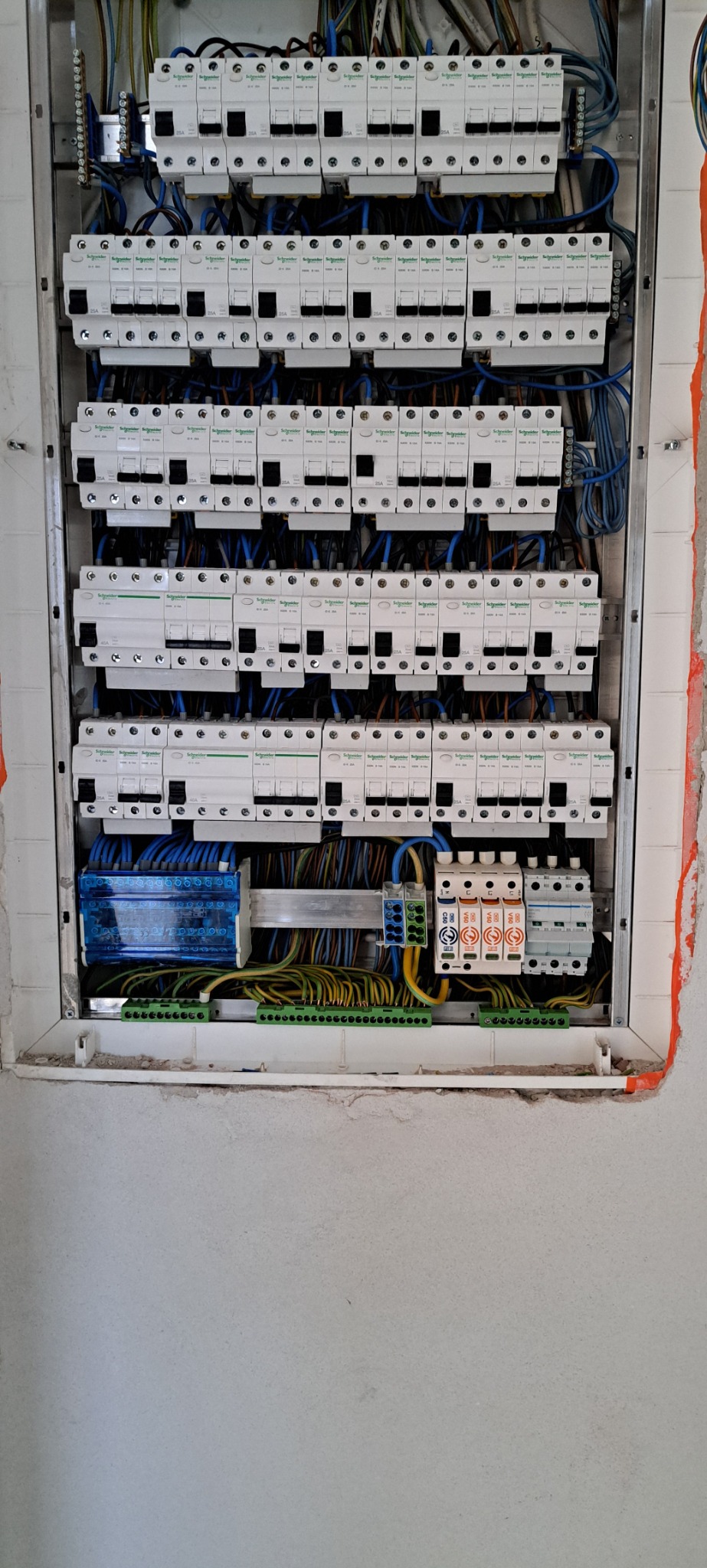 Rozdzielnica elektryczna z zamontowanymi bezpiecznikami i przewodami, widoczne elementy instalacji elektrycznej w trakcie montażu, ściana z widocznymi śladami prac wykończeniowych.