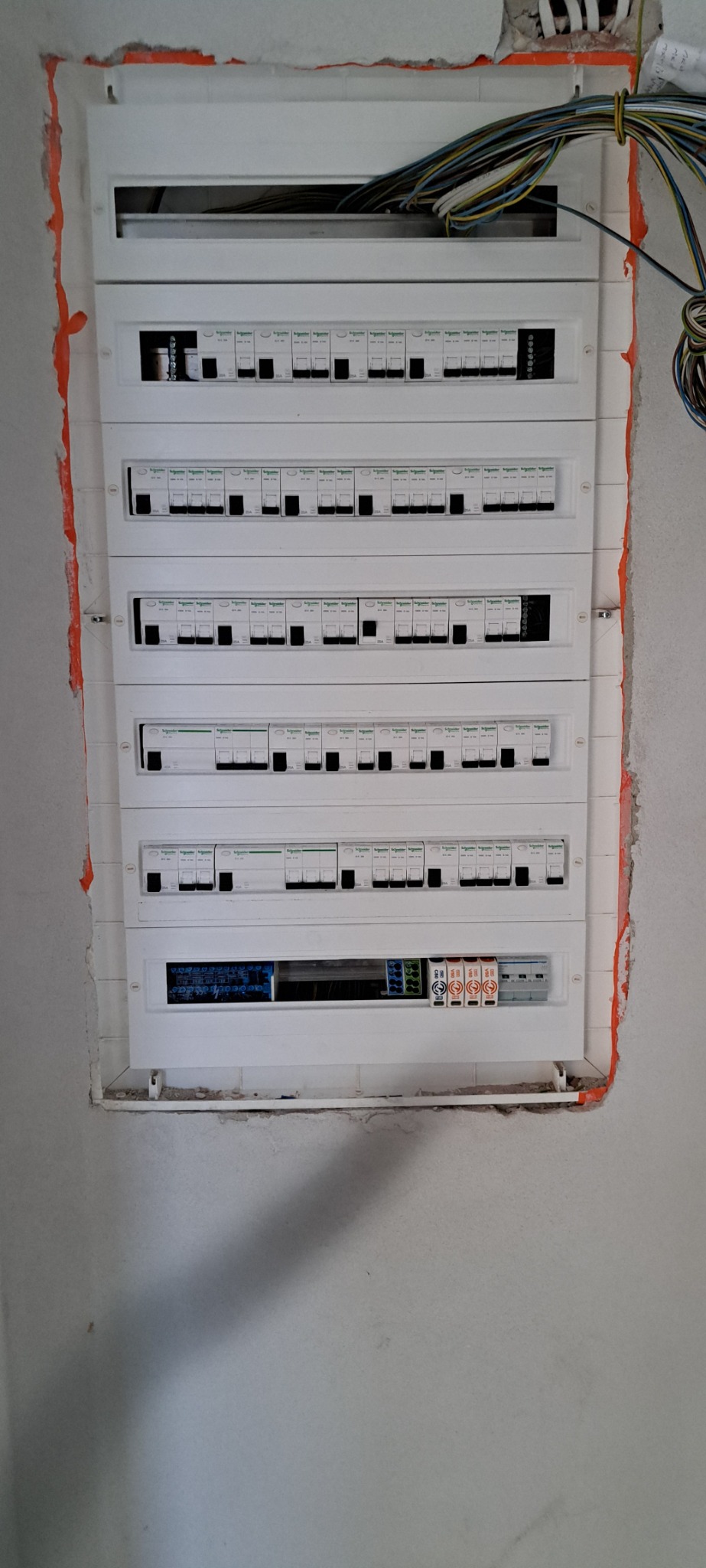 Rozdzielnica elektryczna z widocznymi bezpiecznikami i włącznikami, zamontowana w ścianie w trakcie prac wykończeniowych, z widocznymi przewodami u góry i pomarańczową taśmą zabezpieczającą krawędzie.