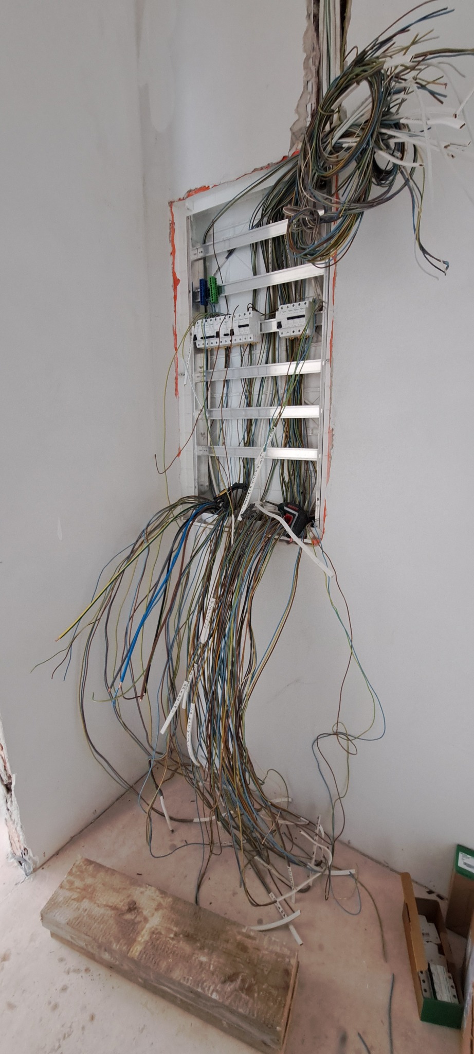 Rozdzielnica elektryczna w trakcie montażu, z licznymi przewodami wychodzącymi ze ściany i częściowo podłączonymi, widoczne zabezpieczenia, skrzynka w surowym stanie, ściany przygotowane...