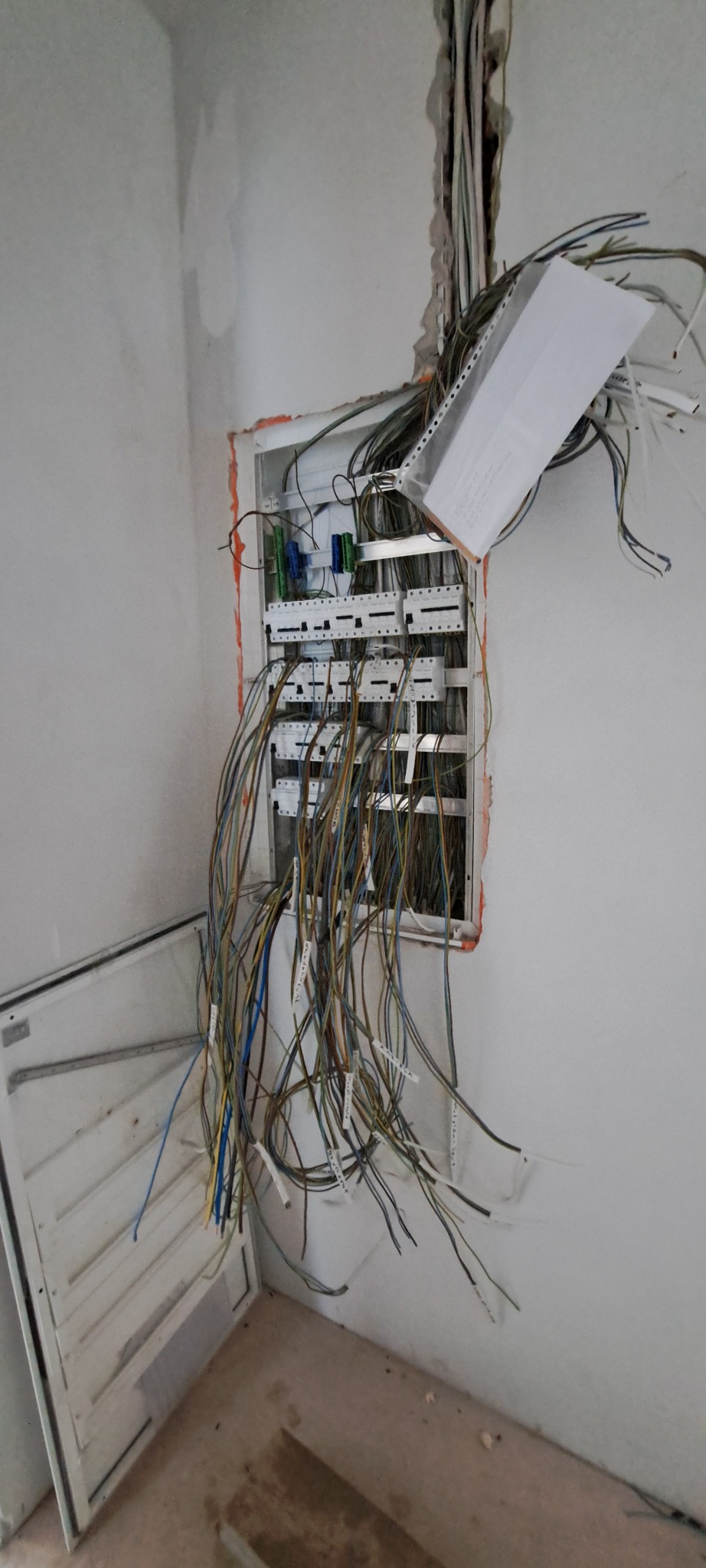 Otwarta skrzynka rozdzielcza z licznymi oznaczonymi przewodami elektrycznymi wychodzącymi na zewnątrz, widoczne elementy montażowe wewnątrz, ściana w stanie surowym.