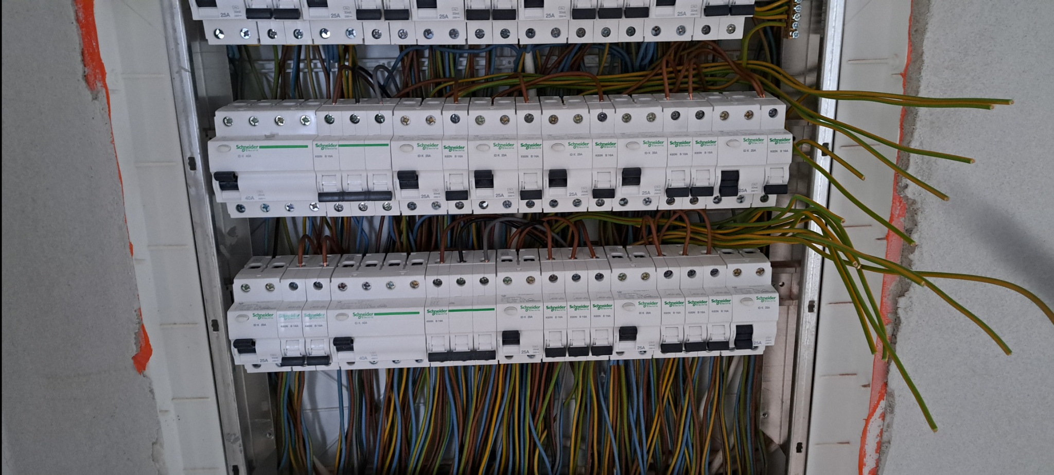 Rozdzielnica elektryczna w trakcie montażu, widoczne zabezpieczenia różnicowoprądowe marki Schneider Electric oraz kable w różnych kolorach, wystające ze ściany, z fragmentami pomarańczowej farby...