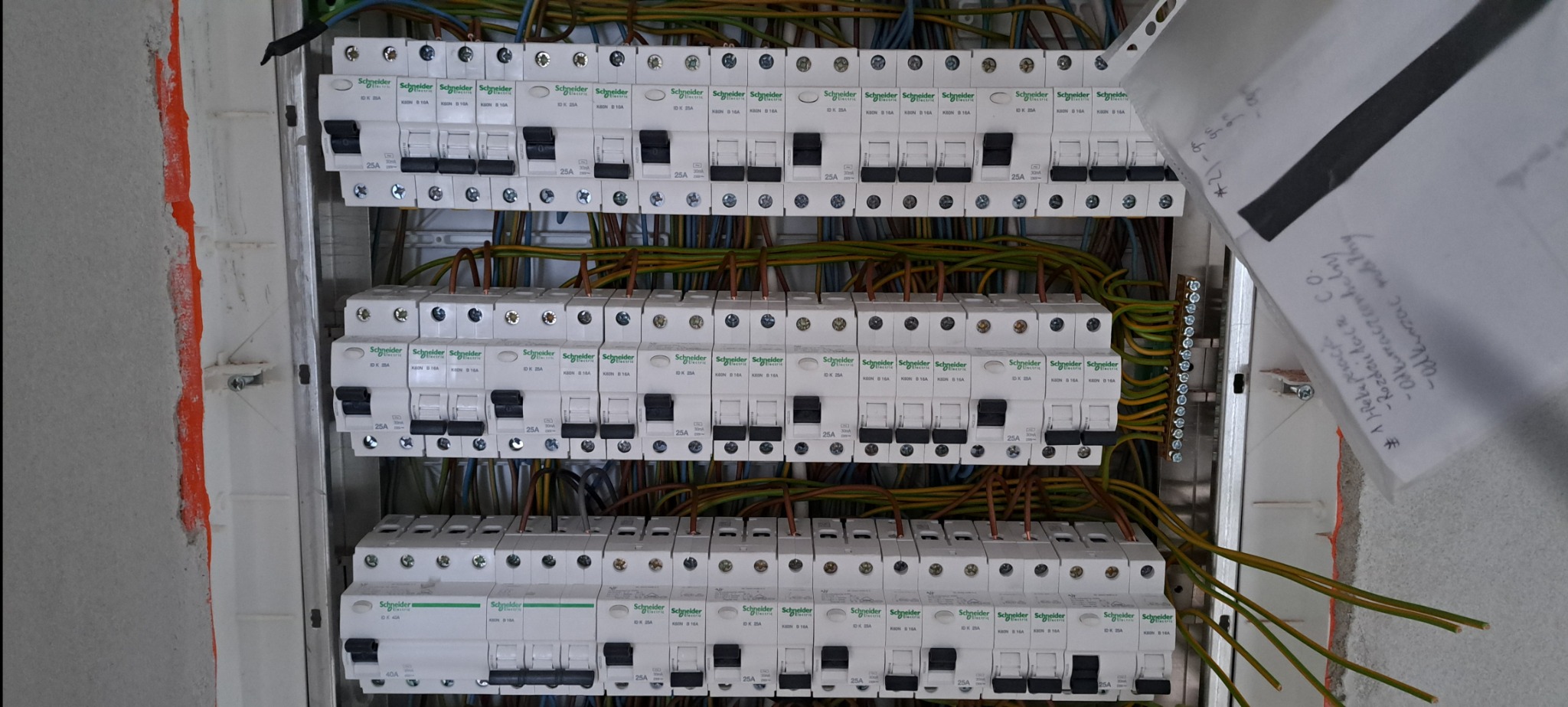 Rozdzielnica elektryczna z zamontowanymi bezpiecznikami Schneider Electric, widoczne kable i przewody, obok rozdzielnicy kartka z notatkami.