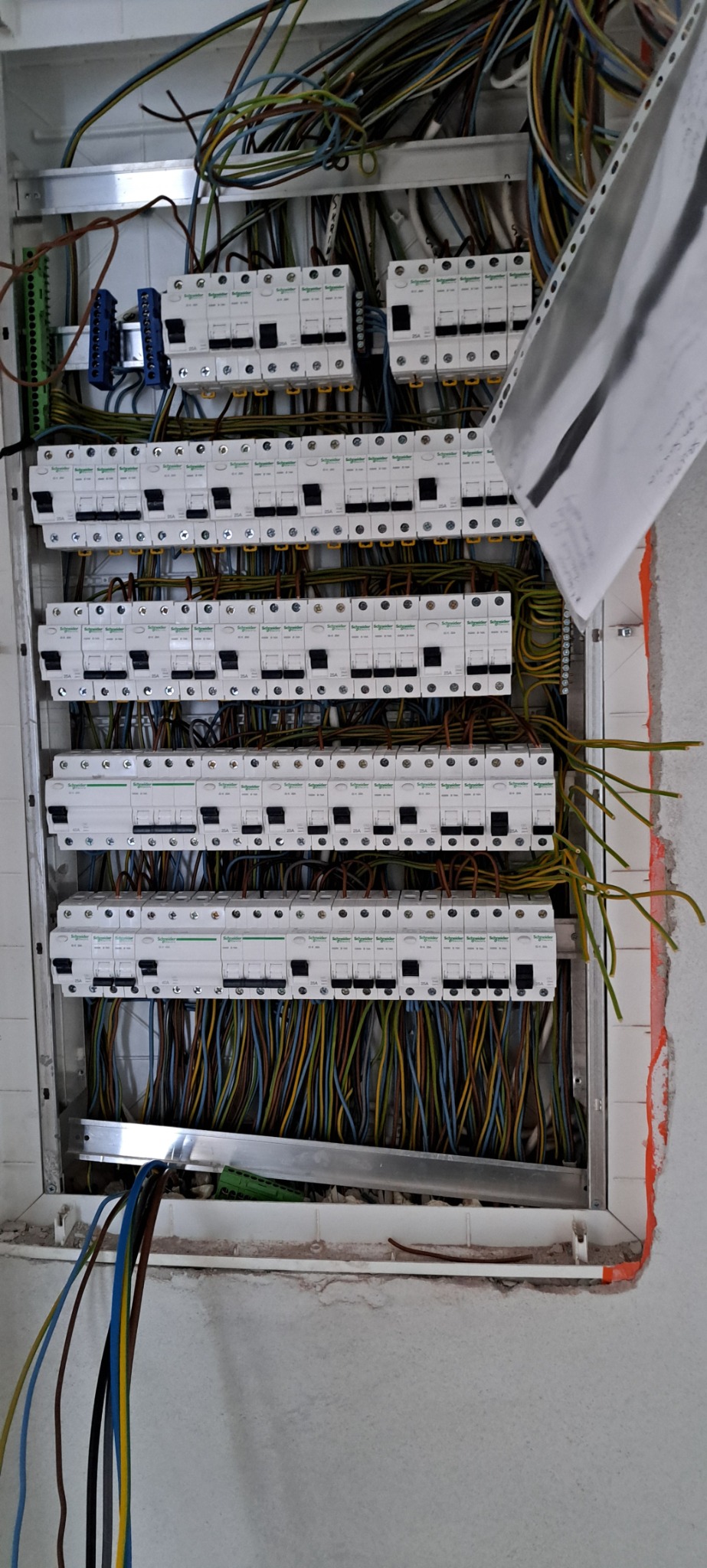 Otwarta skrzynka elektryczna z zamontowanymi bezpiecznikami Schneider Electric i licznymi przewodami w różnych kolorach, widoczne także fragmenty dokumentacji technicznej.