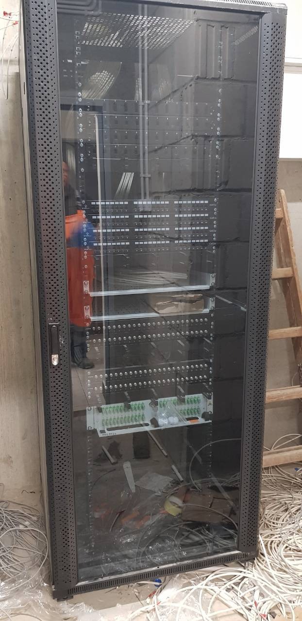 Szafa rack z zamontowanymi modułami i panelami krosowniczymi, widoczne liczne przewody wewnątrz i na podłodze, drabina oparta o ścianę w tle.
