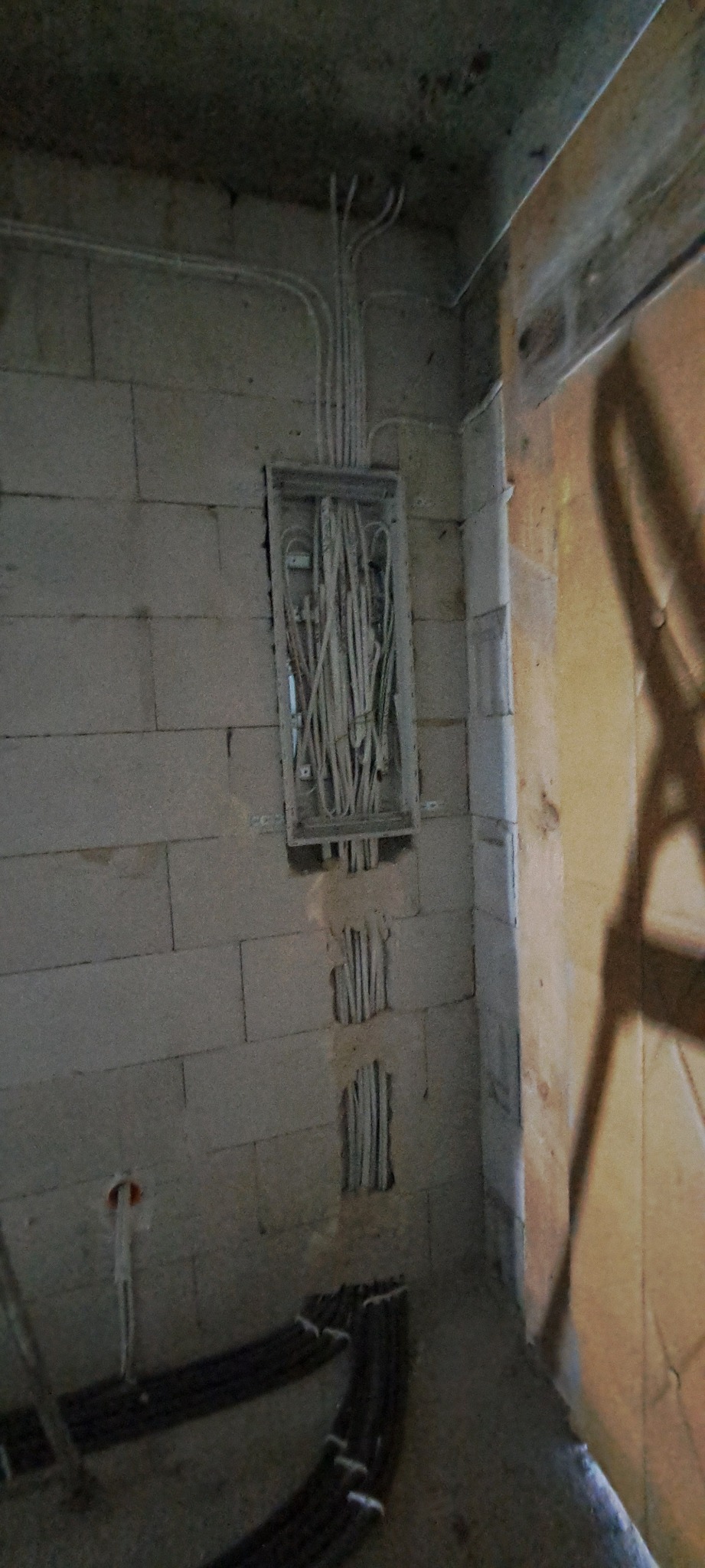 Rozdzielnica elektryczna w stanie surowym, widoczne przewody w puszce i prowadzone w bruzdach w ścianie z bloczków, fragment drabiny malarskiej w tle.