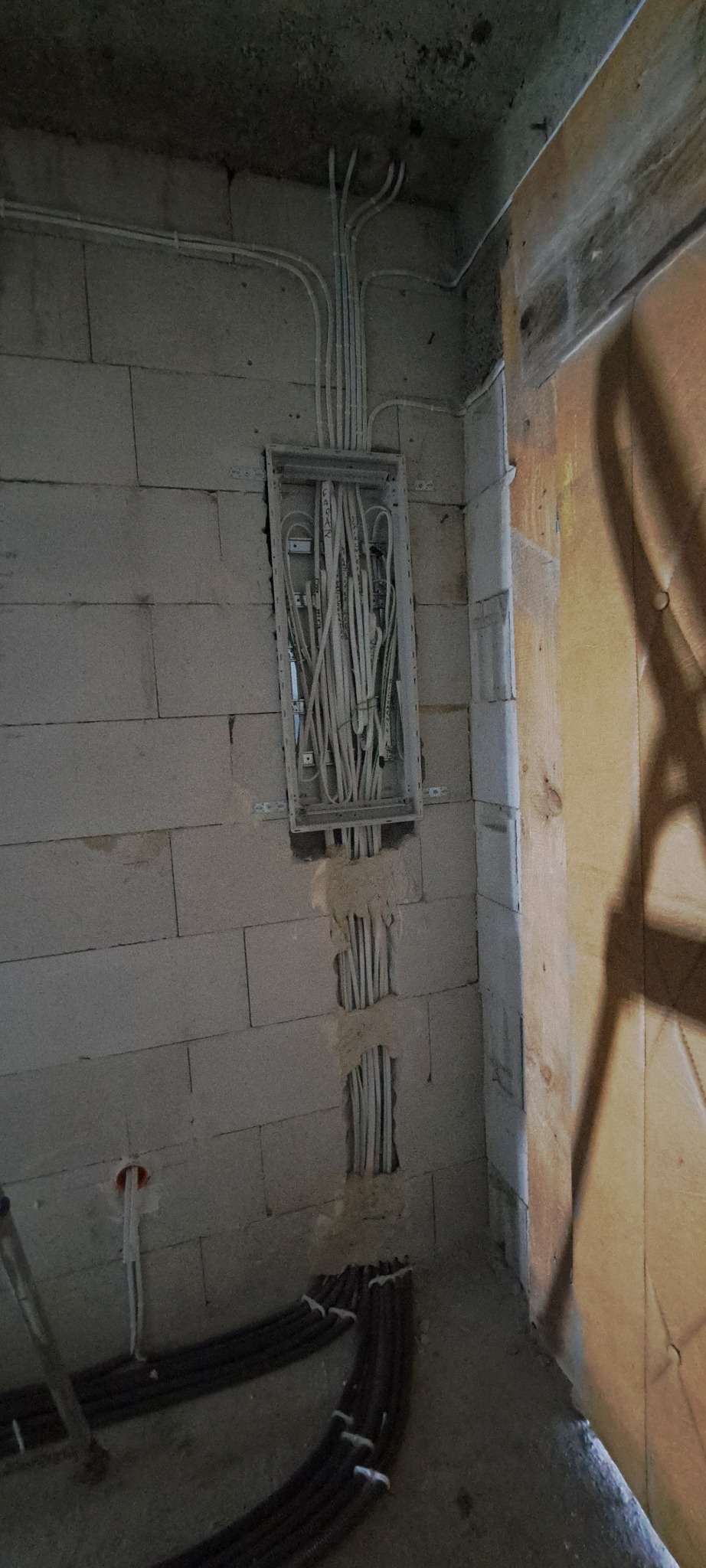 Rozdzielnica elektryczna z poprowadzonymi przewodami w surowym pomieszczeniu z bloczków, widoczne kable w ścianie i na podłodze w peszlach.