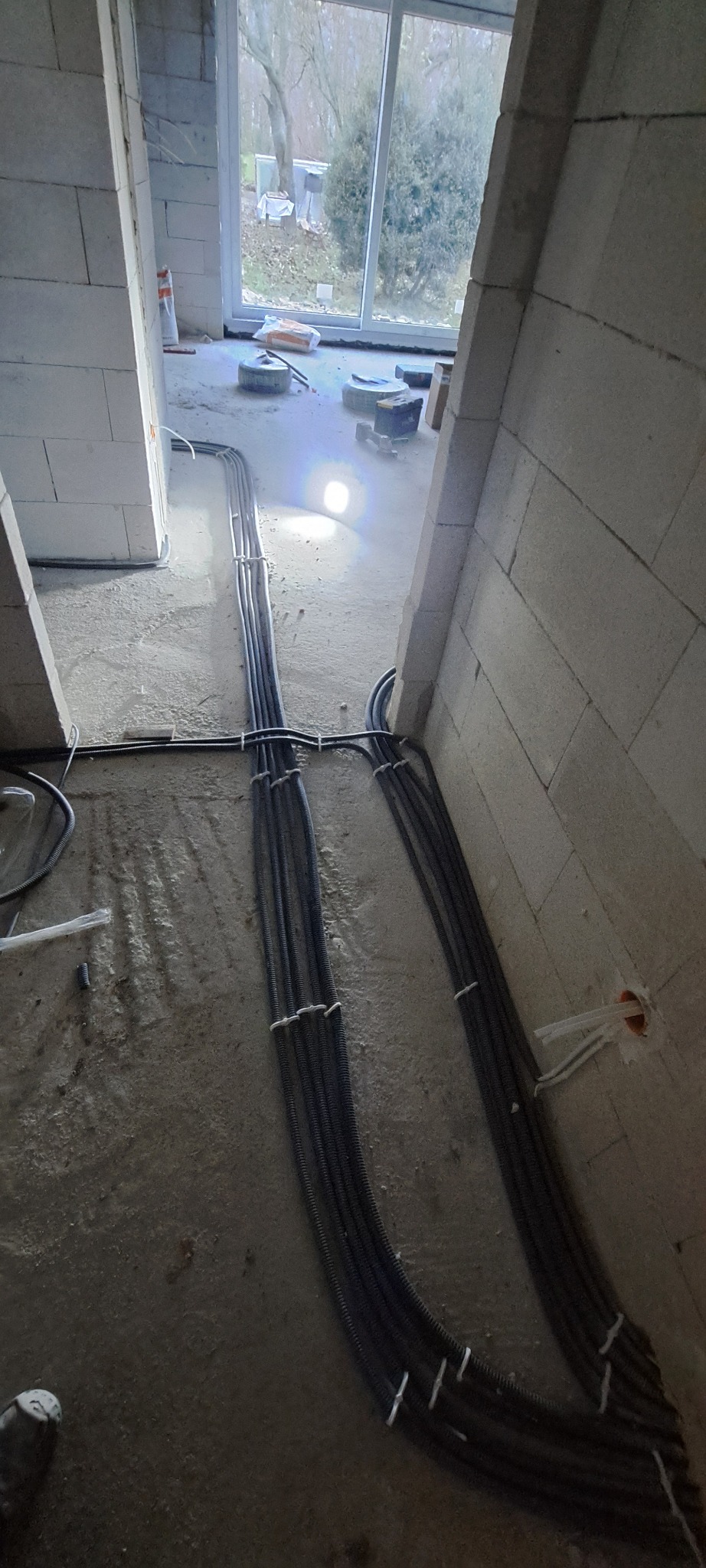 Wiązki przewodów elektrycznych w peszlach ułożone na surowej podłodze w trakcie instalacji w nowym budynku z pustaków, widoczne okno i materiały budowlane w tle.
