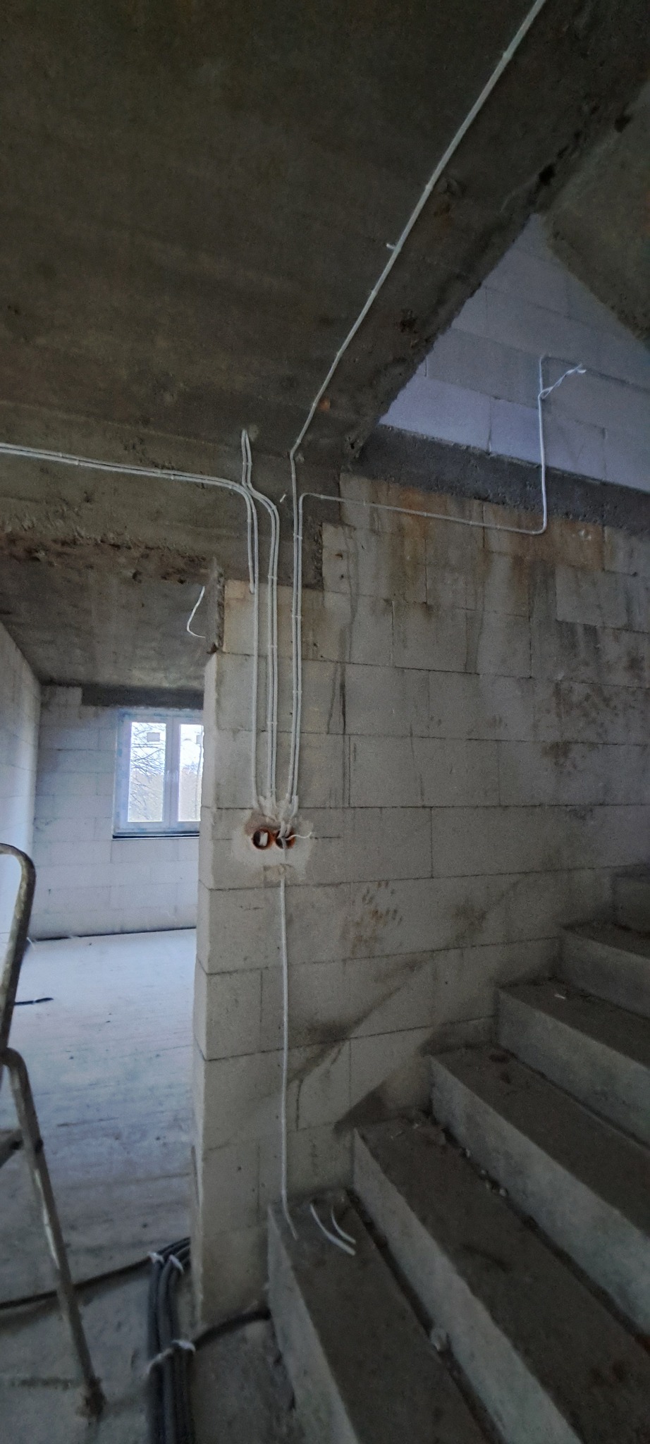 Surowy stan budynku w trakcie prac elektrycznych: kable prowadzone po ścianach z bloków, widoczne puszki instalacyjne, fragment schodów i drabina w tle.
