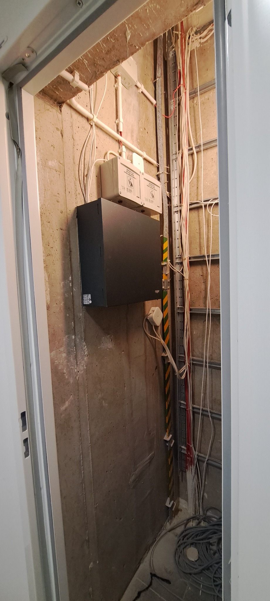 Węzeł instalacyjny w surowym pomieszczeniu, widoczne przewody elektryczne, puszki przyłączeniowe, metalowa szyna montażowa, czarna skrzynka Roger, oraz zwój kabli na podłodze.