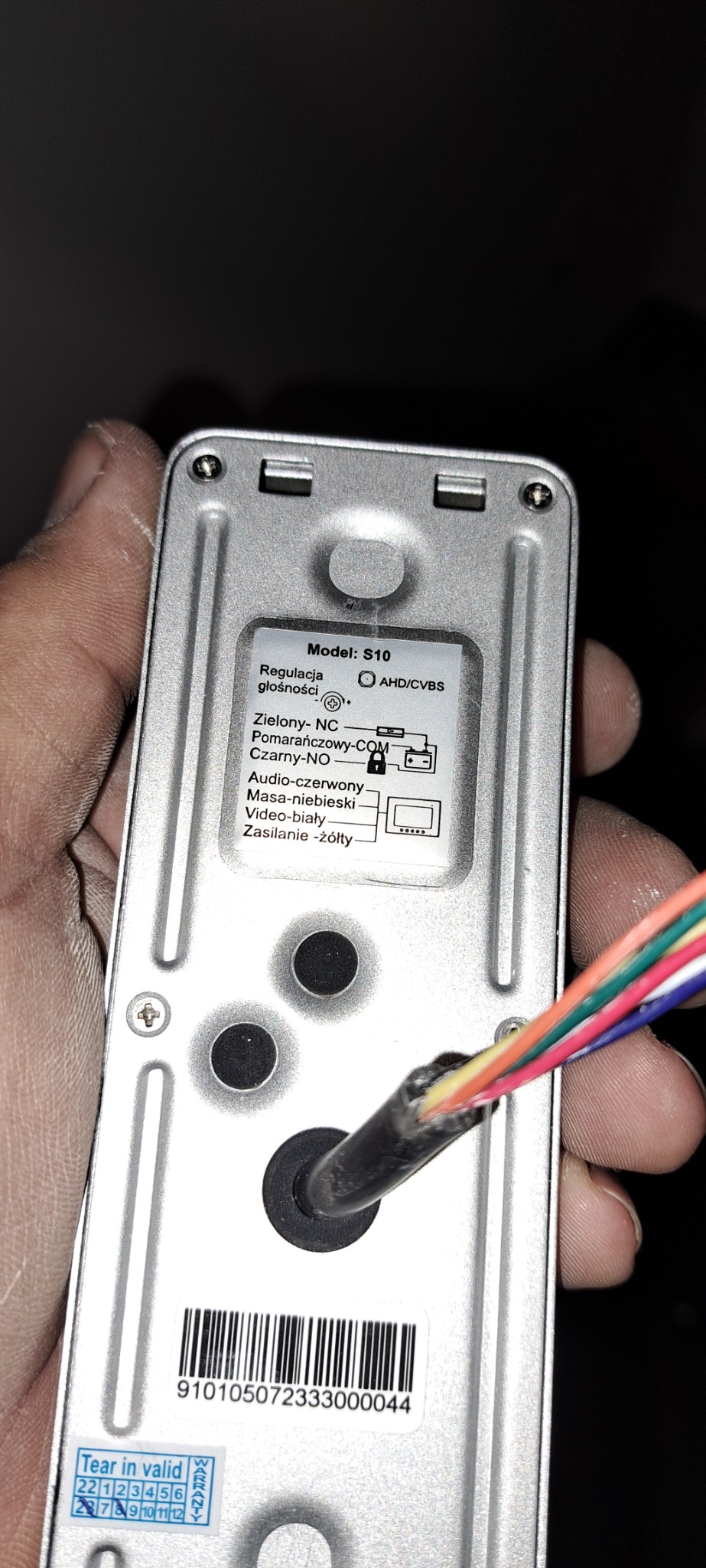 Tył obudowy wideodomofonu S10 z widoczną tabliczką z opisem podłączeń, trzymany w dłoni, z wychodzącym kablem wielożyłowym.