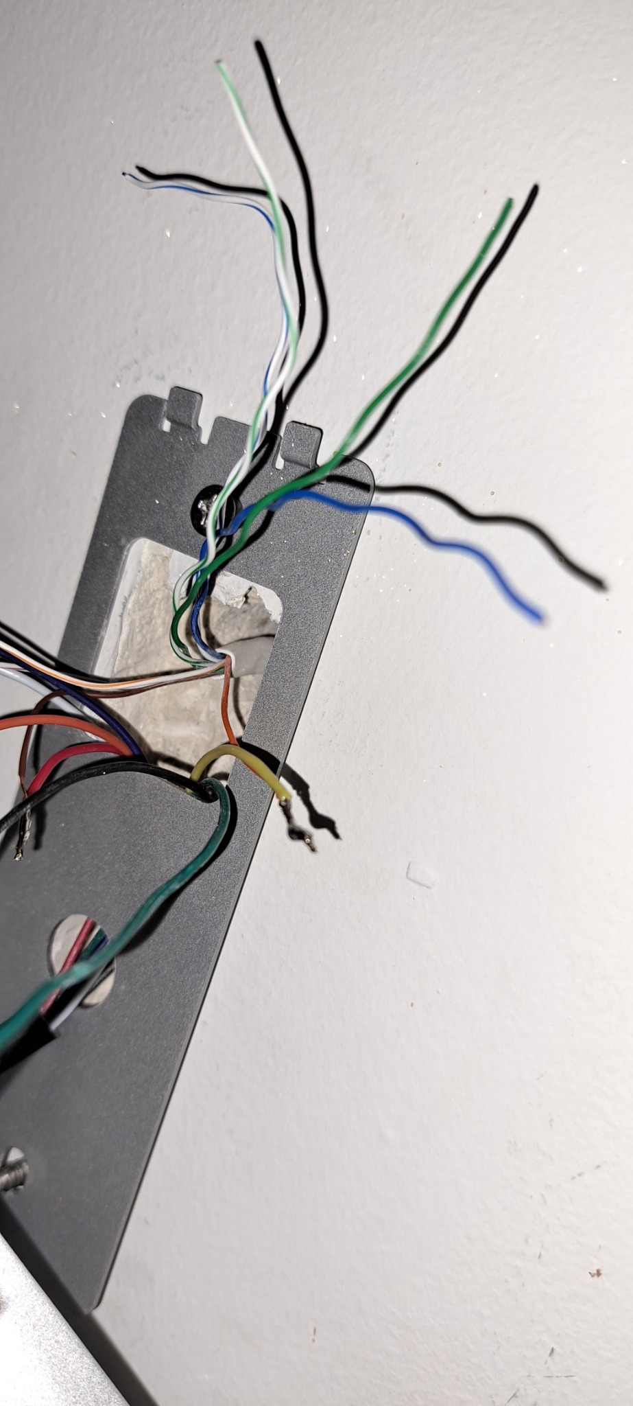 Metalowa płytka montażowa z wystającymi przewodami o różnych kolorach (zielony, niebieski, biały, czarny, czerwony, żółty) przymocowana do białej ściany. Widoczne ślady po wierceniu w ścianie.