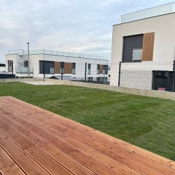 Kamil Łakomy - Nowoczesny ogród na dachu z drewnianym tarasem i świeżą trawą, na tle budynków z metalowymi schodami spiralnymi.