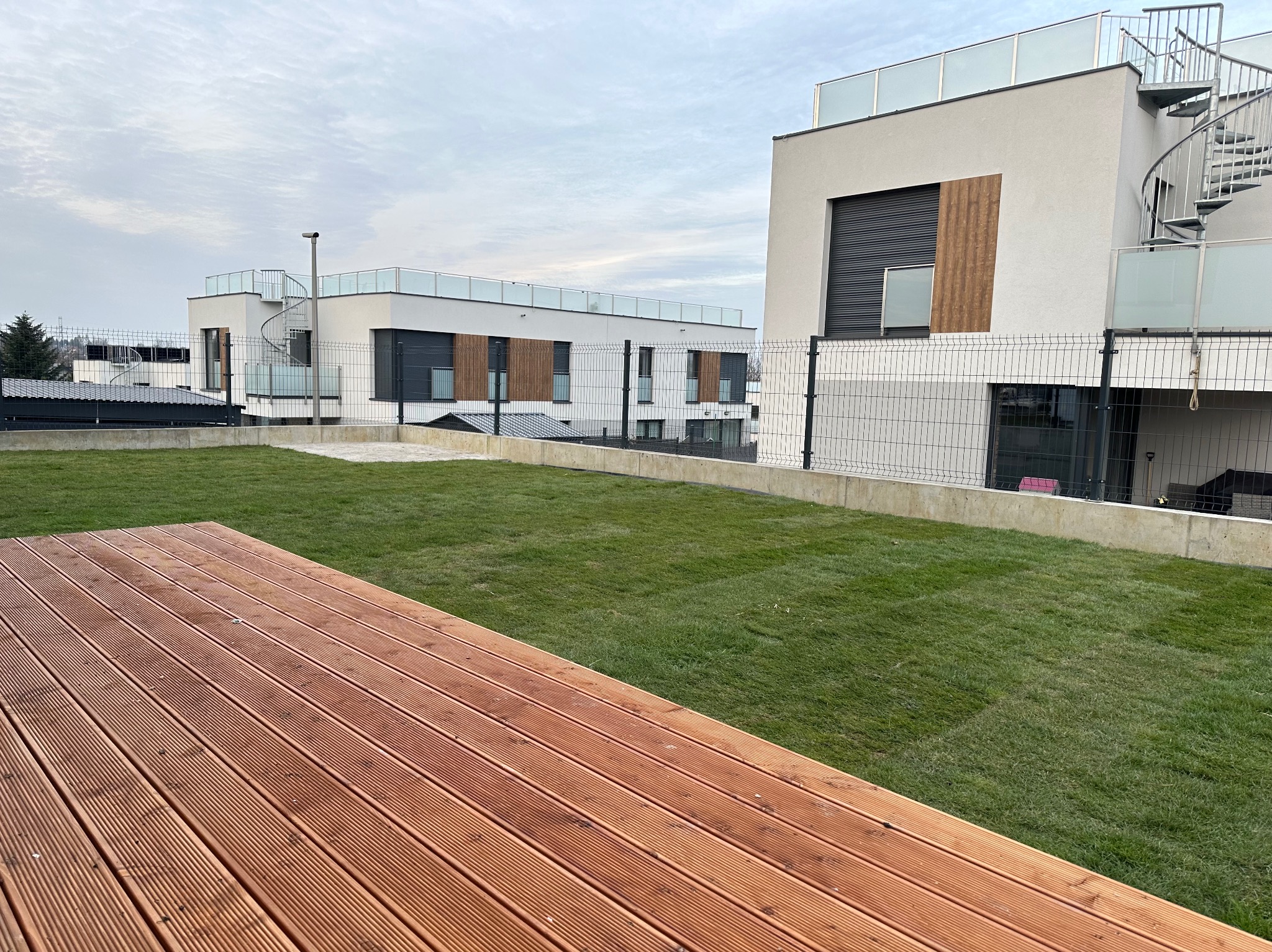 Nowoczesny ogród na dachu z drewnianym tarasem i świeżą trawą, na tle budynków z metalowymi schodami spiralnymi.