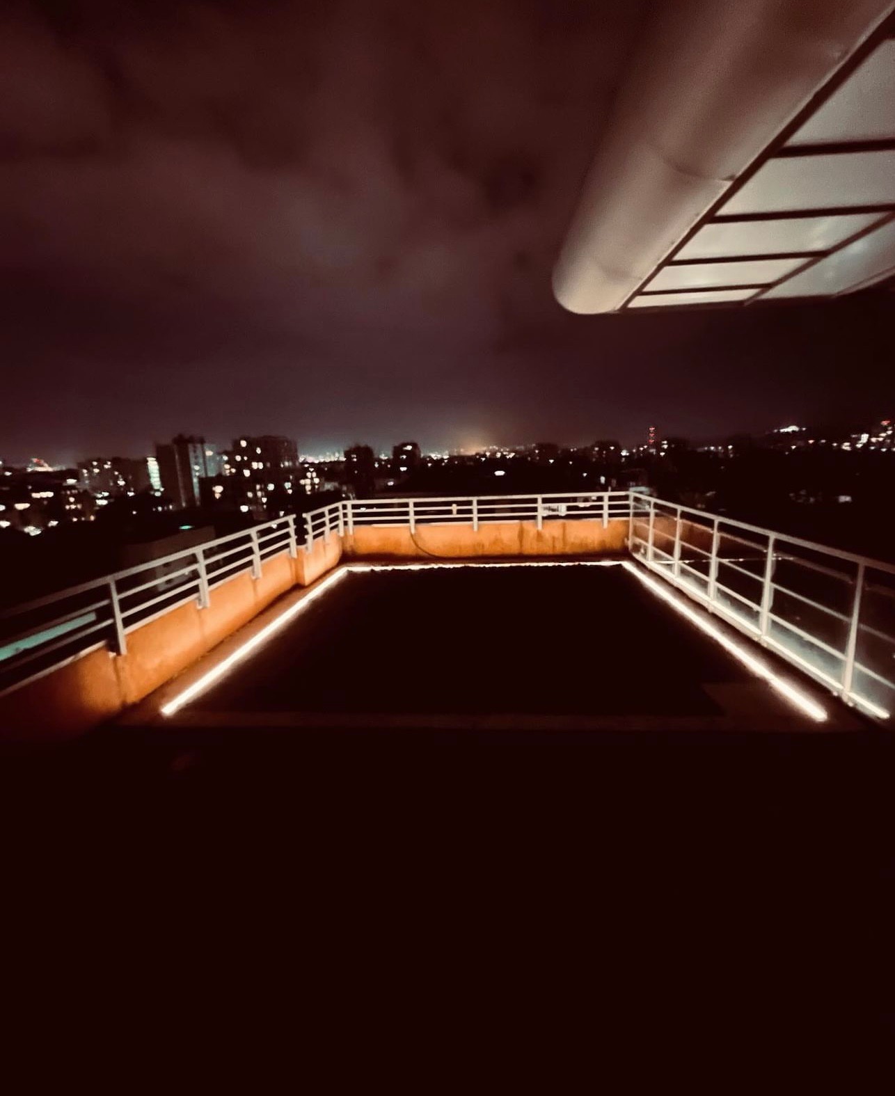 Nocny widok z tarasu na dachu budynku z oświetleniem LED wzdłuż krawędzi i panoramą miasta w tle pod ciemnym, zachmurzonym niebem.