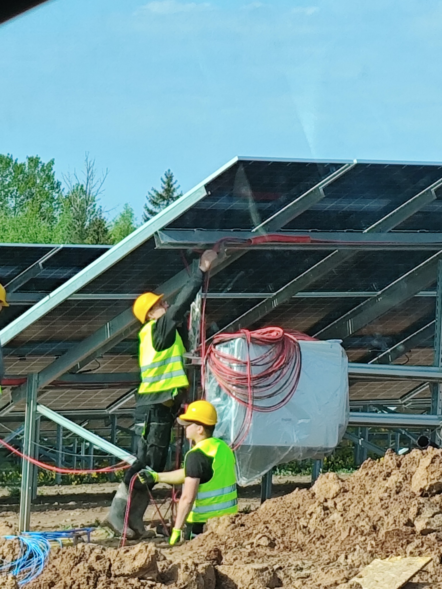 Montaż paneli słonecznych: dwóch instalatorów w kaskach i kamizelkach łączy okablowanie na konstrukcji fotowoltaicznej w słoneczny dzień, widoczne drzewa w tle.