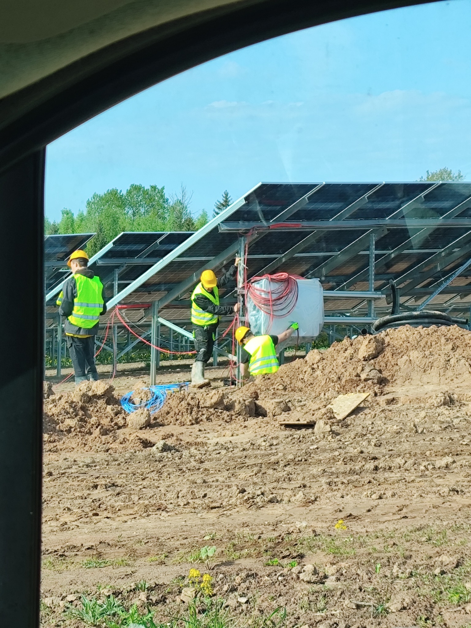 Montaż paneli słonecznych: trzech monterów w żółtych kaskach instaluje system fotowoltaiczny na polu. Widoczne kable i elementy konstrukcyjne.