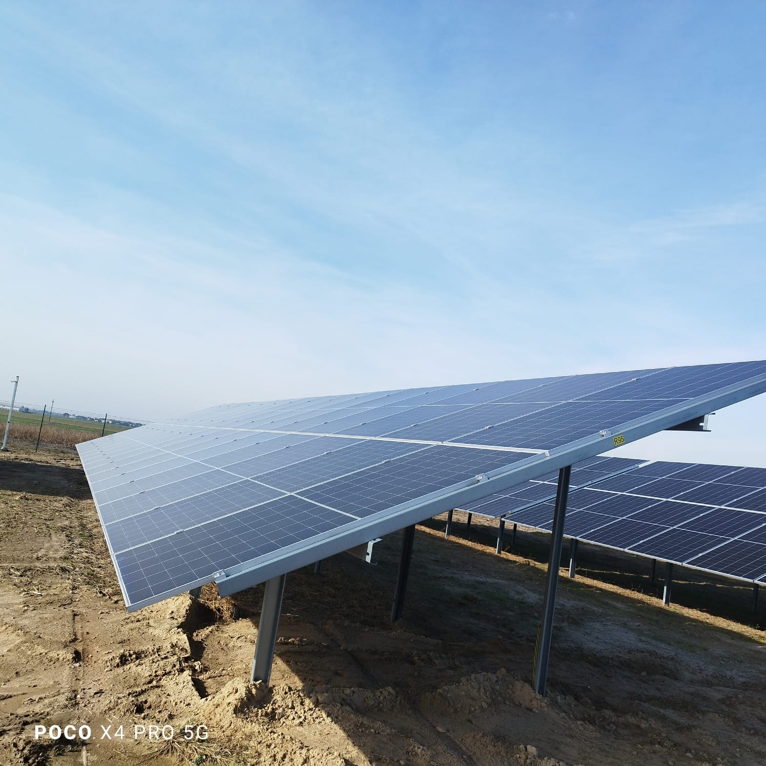 Rząd paneli fotowoltaicznych na piaszczystym gruncie pod błękitnym niebem. Widoczne metalowe konstrukcje wsporcze i kolejne rzędy paneli w oddali.