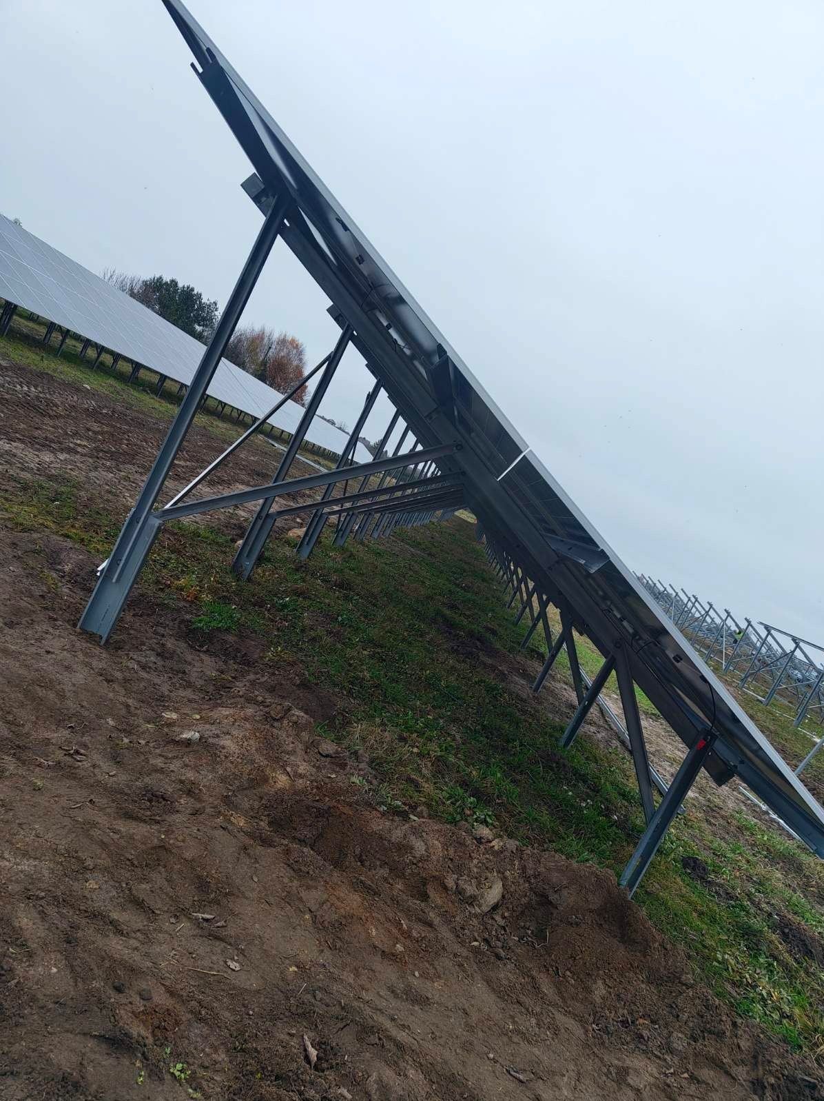 Nisko kątowe ujęcie rzędu paneli fotowoltaicznych z metalową konstrukcją na nierównym, trawiastym terenie w pochmurny dzień. Widoczne kolejne rzędy paneli w tle.