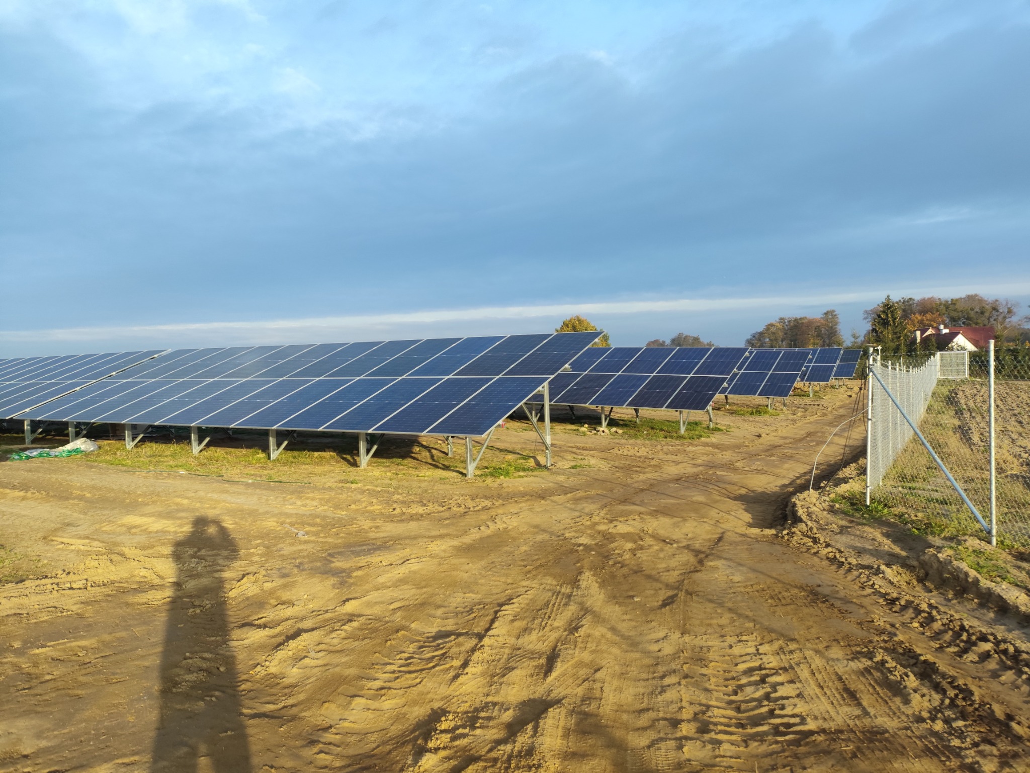Rozległa instalacja paneli słonecznych na ziemnym podłożu, z widocznym cieniem osoby fotografującej, ogrodzeniem i budynkiem w oddali pod błękitnym niebem.