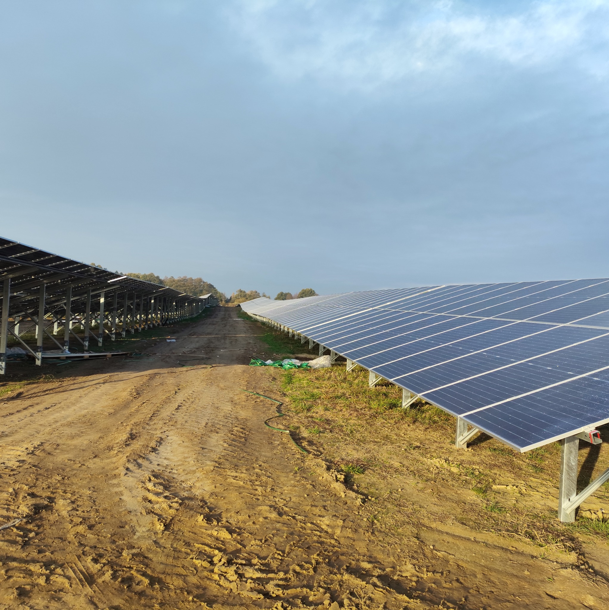 Rozległa farma paneli słonecznych w polu, z ziemistą drogą w perspektywie i pochmurnym niebem. Panele ustawione w rzędach, widoczne ślady opon.