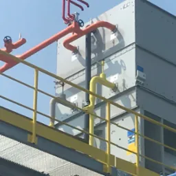 Widok z dołu na przemysłową konstrukcję z metalowymi rurami w kolorach pomarańczowym i żółtym, połączonymi z szarą, modułową konstrukcją, prawdopodobnie chłodniczą, z widocznym logo BAC.