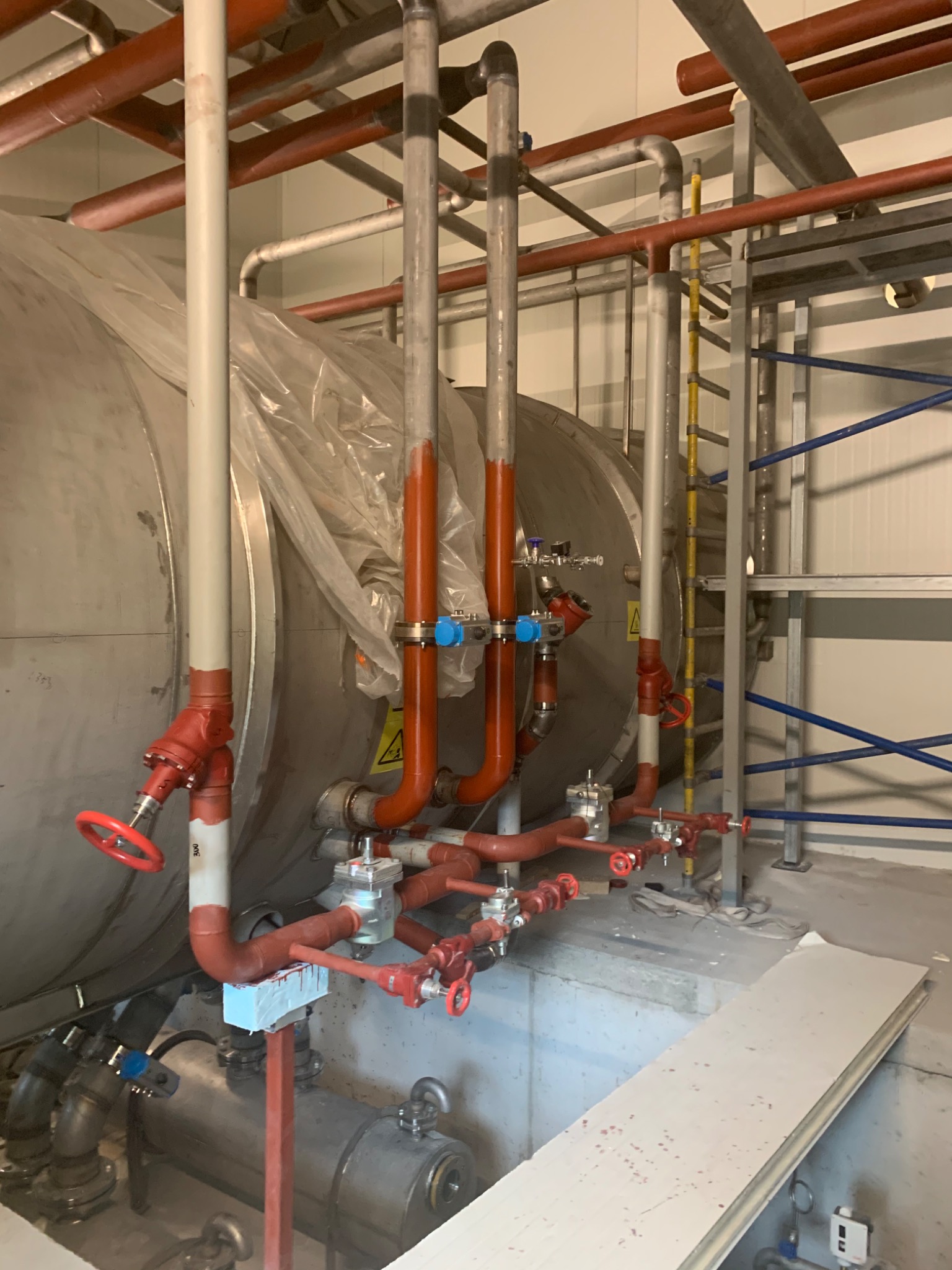 Srebrny zbiornik przemysłowy z licznymi rurami, zaworami i osprzętem, niektóre rury pomalowane na czerwono, widoczne znaki ostrzegawcze, częściowo owinięty folią ochronną.