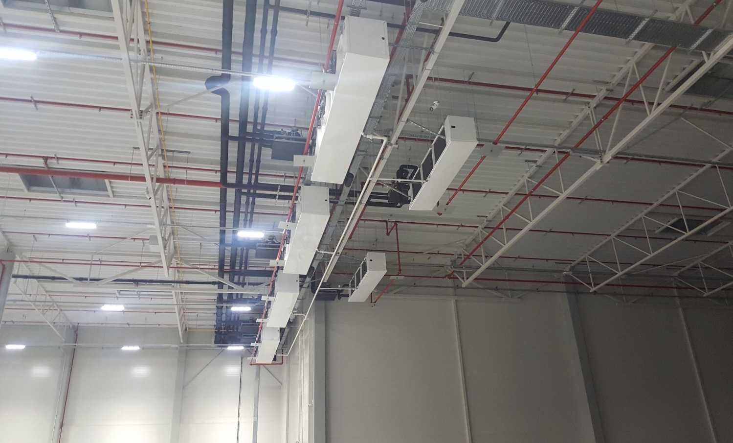 Wnętrze hali produkcyjnej z widoczną konstrukcją stalową dachu, instalacjami wentylacyjnymi i rurociągami.