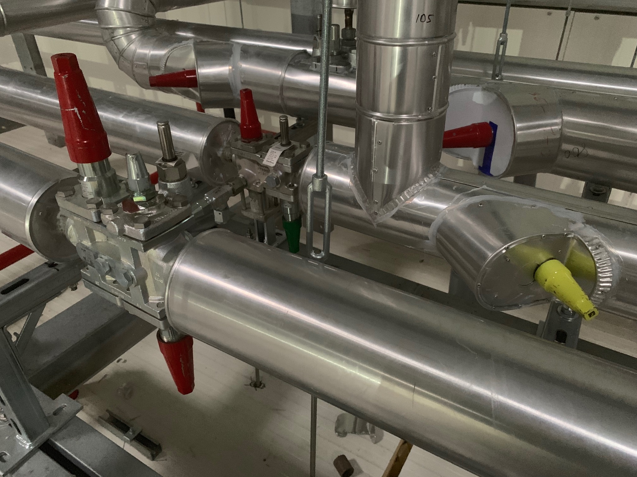 Instalacja przemysłowa z rurami ze stali nierdzewnej, zaworami z czerwonymi uchwytami i wspornikami montażowymi, widoczne oznaczenia '105' na jednej z rur.