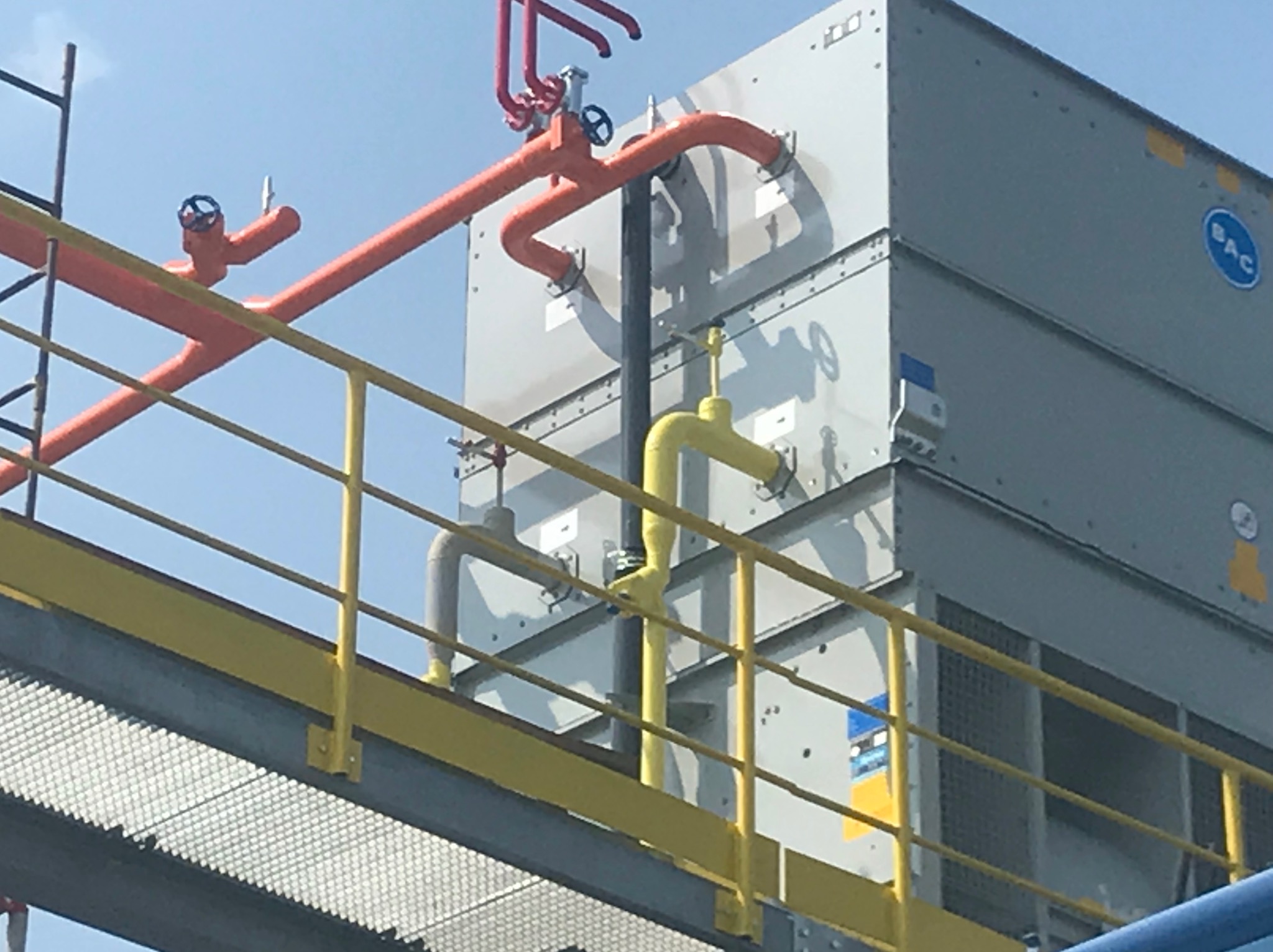 Widok z dołu na przemysłową konstrukcję z metalowymi rurami w kolorach pomarańczowym i żółtym, połączonymi z szarą, modułową konstrukcją, prawdopodobnie chłodniczą, z widocznym logo BAC.