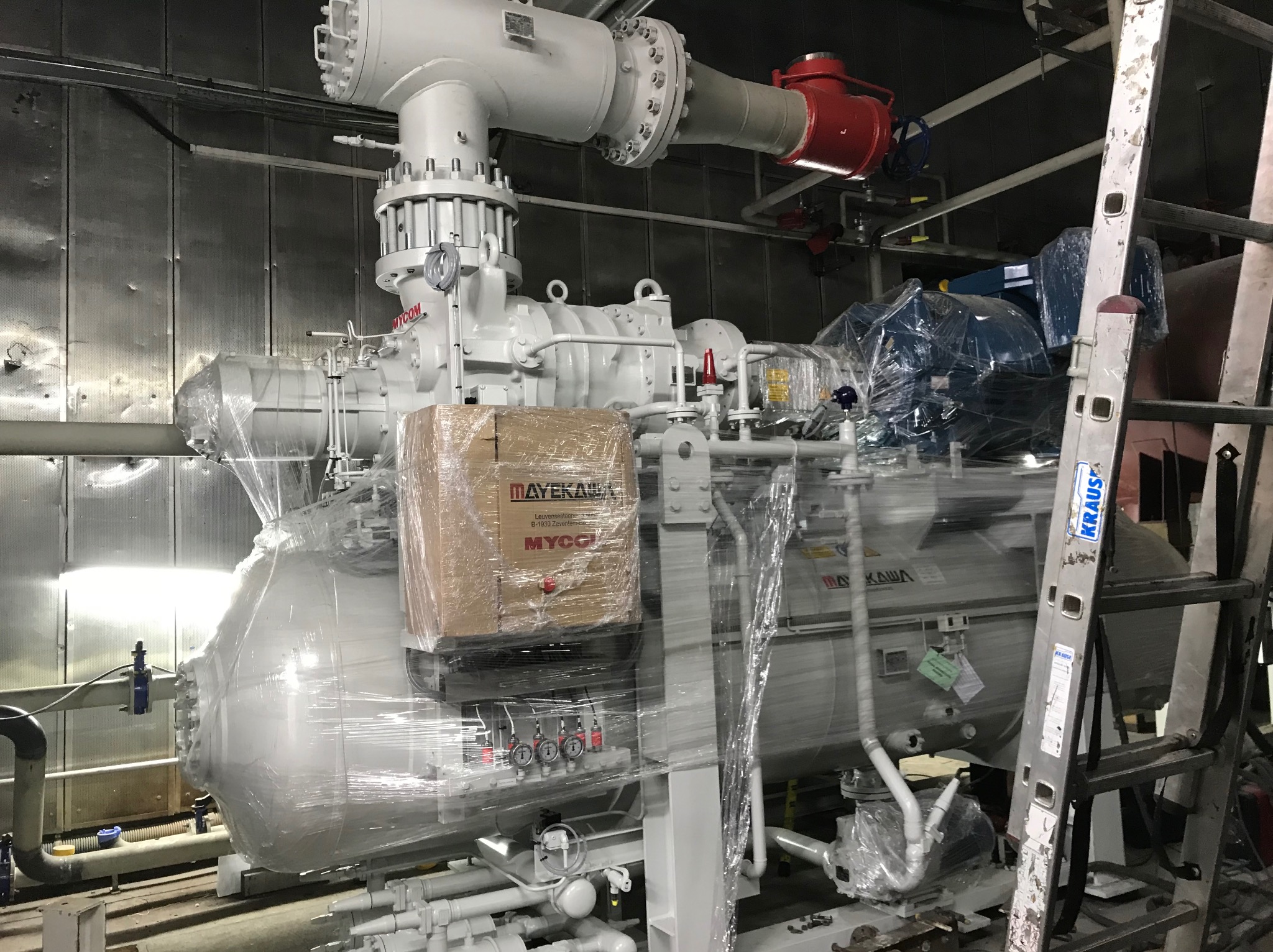 Nowa, biała przemysłowa chłodnica Mayekawa MYCOM, owinięta folią ochronną i zabezpieczona kartonem, umieszczona w pomieszczeniu z metalowymi ścianami, widoczna drabina Kraus.