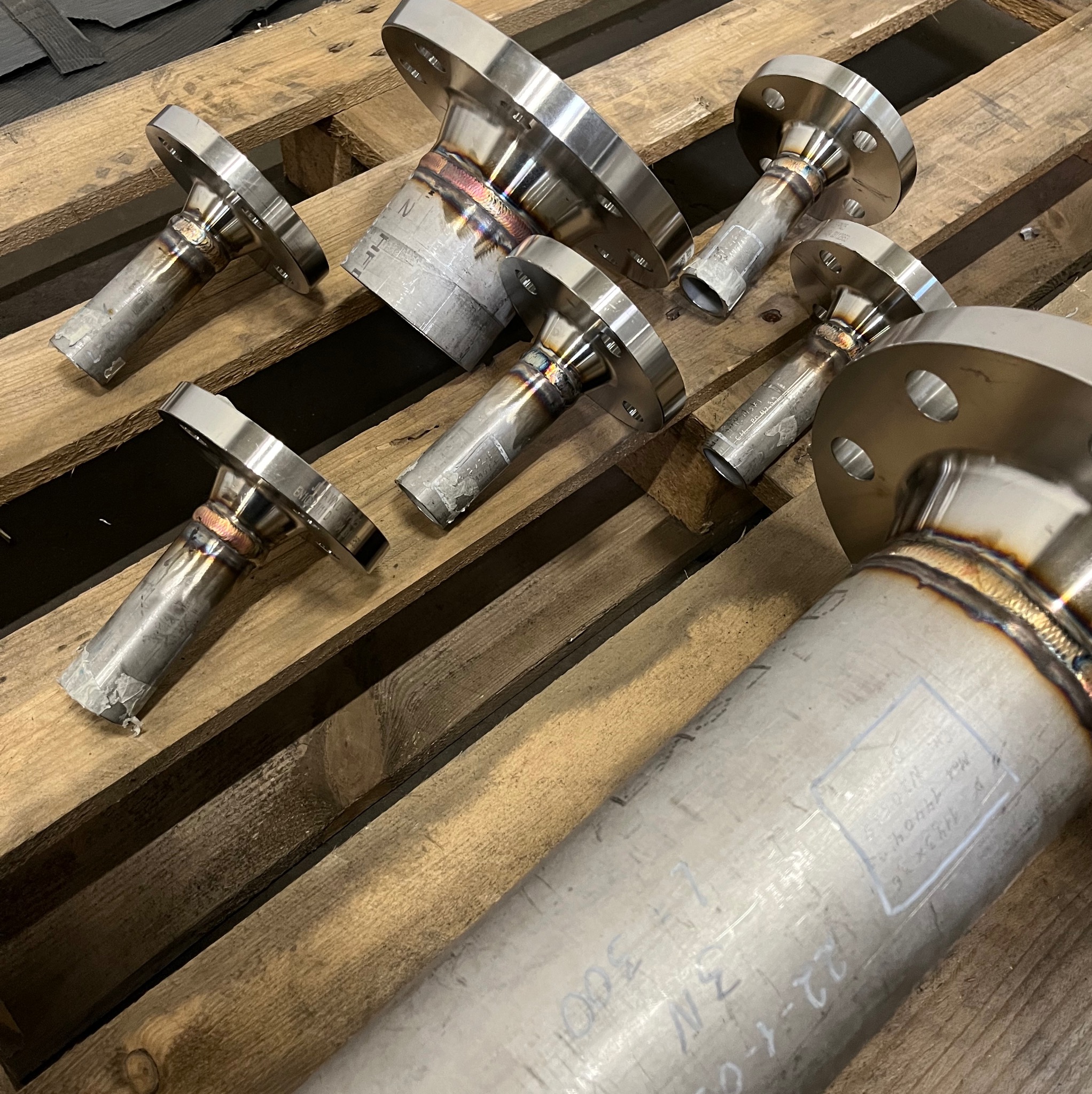 Sześć spawanych, nierdzewnych rur z kołnierzami ułożonych na drewnianej palecie, widoczne ślady po spawaniu i oznaczenia na powierzchni metalu.