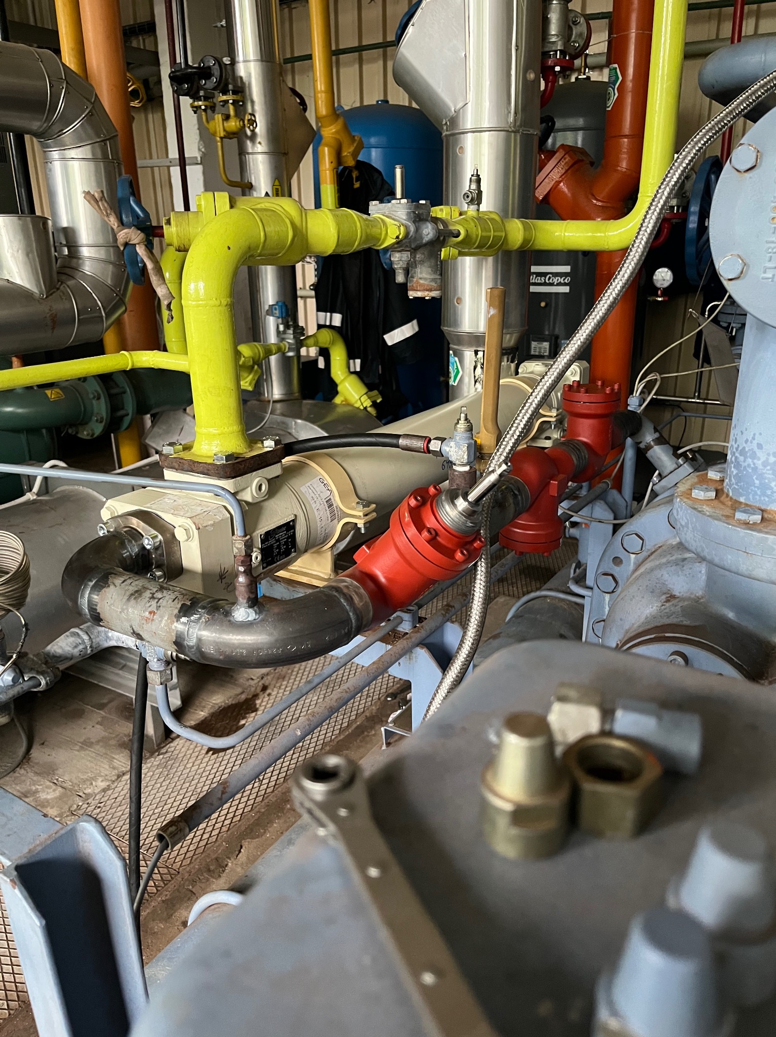 Złożony układ rur i zaworów w hali produkcyjnej, widoczne elementy metalowe, żółte rury, czerwone zawory oraz srebrne przewody elastyczne, w tle maszyny i instalacje przemysłowe.