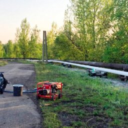 AGWA Sp. z o.o. - Malowanie rurociągu nadziemnego, widoczny agregat malarski, generator prądu i dwóch mężczyzn podczas pracy.